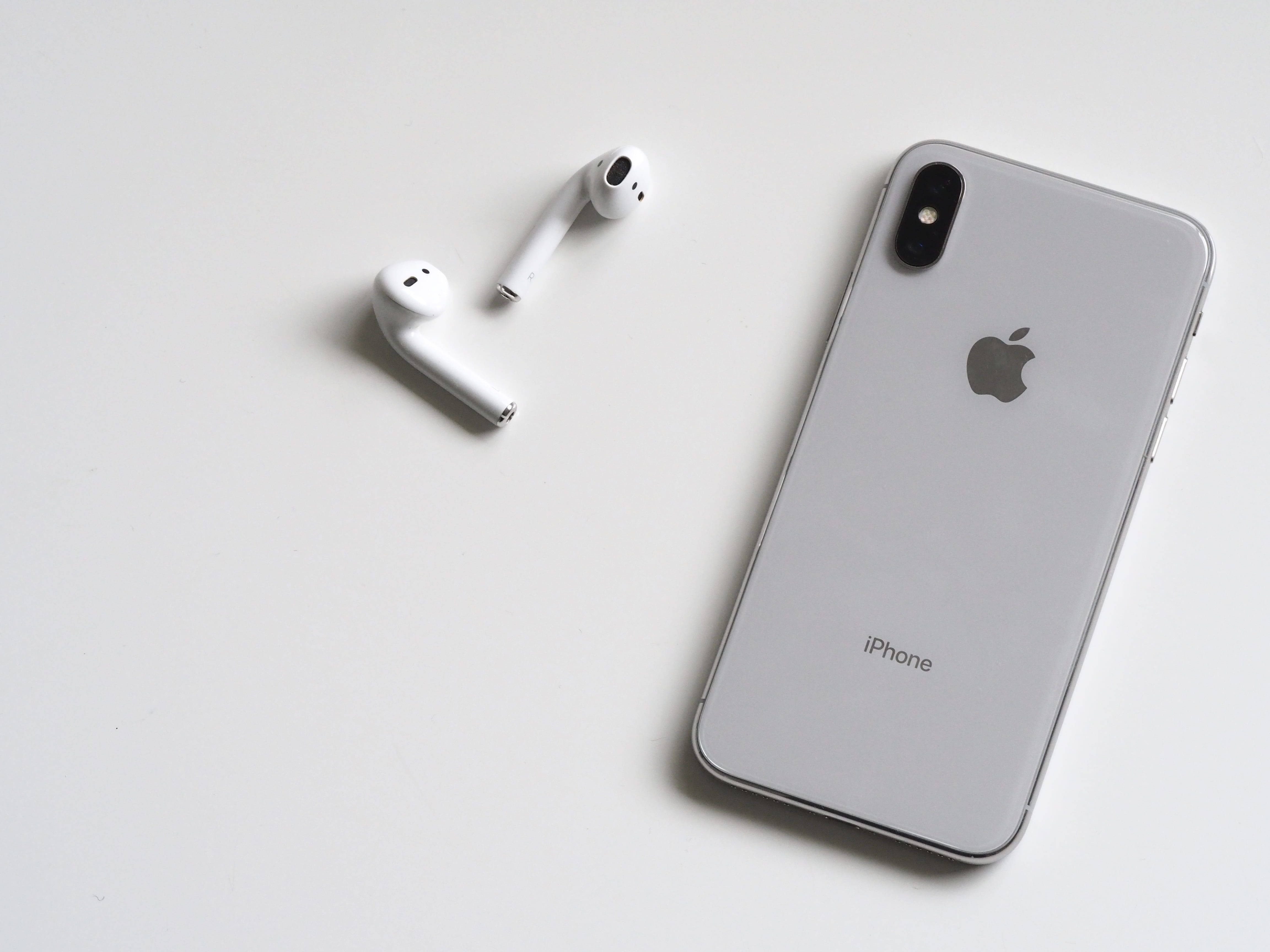 Airpods 桌布 壁紙 — 一部時尚的銀色iPhone X放在其白色無線耳機旁邊，置於簡約的白色表面上。