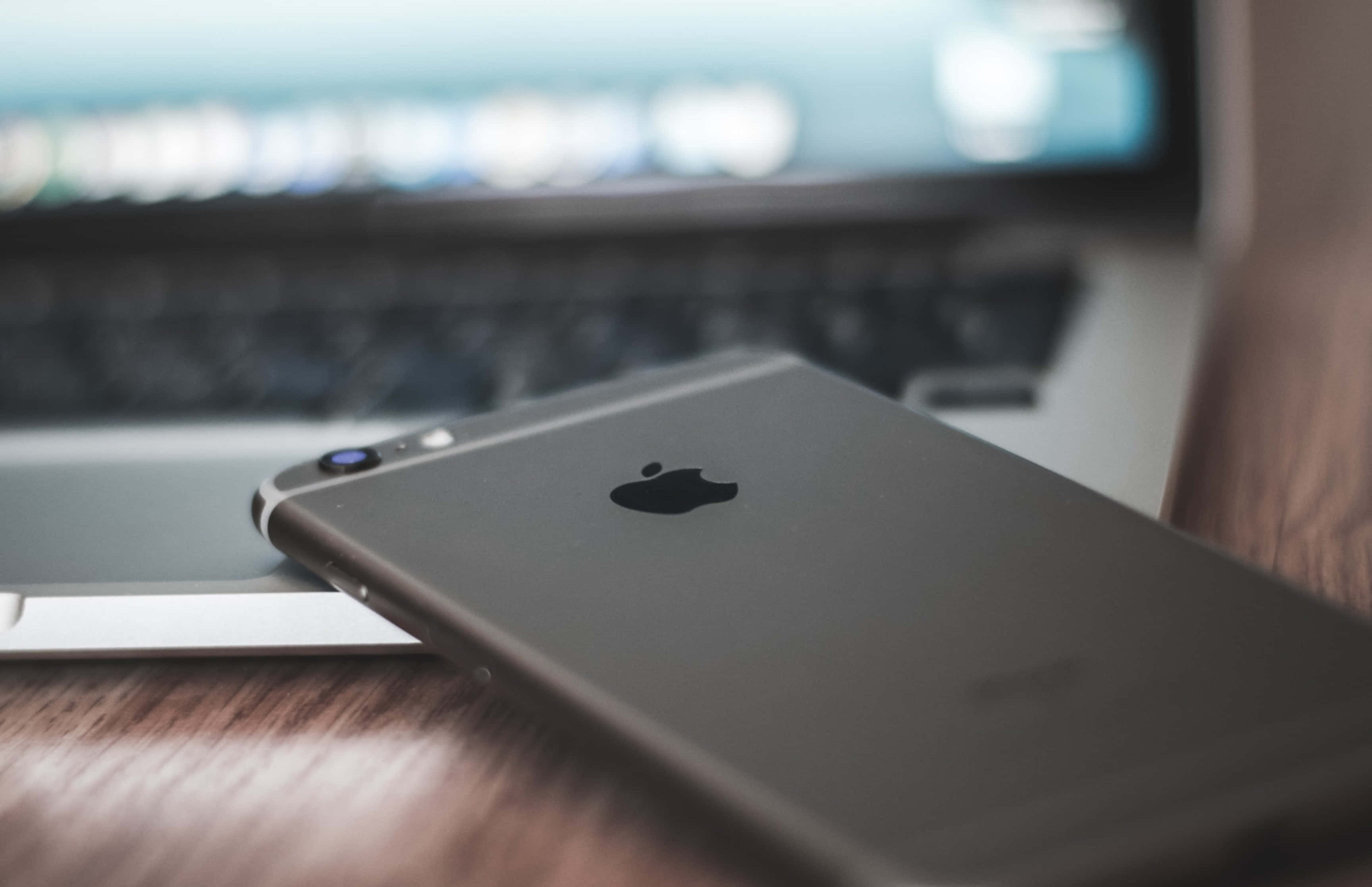 macbook pro 桌布 壁紙 — 一部時尚的銀色iPhone放在木桌上，旁邊是模糊的筆記型電腦鍵盤和螢幕。