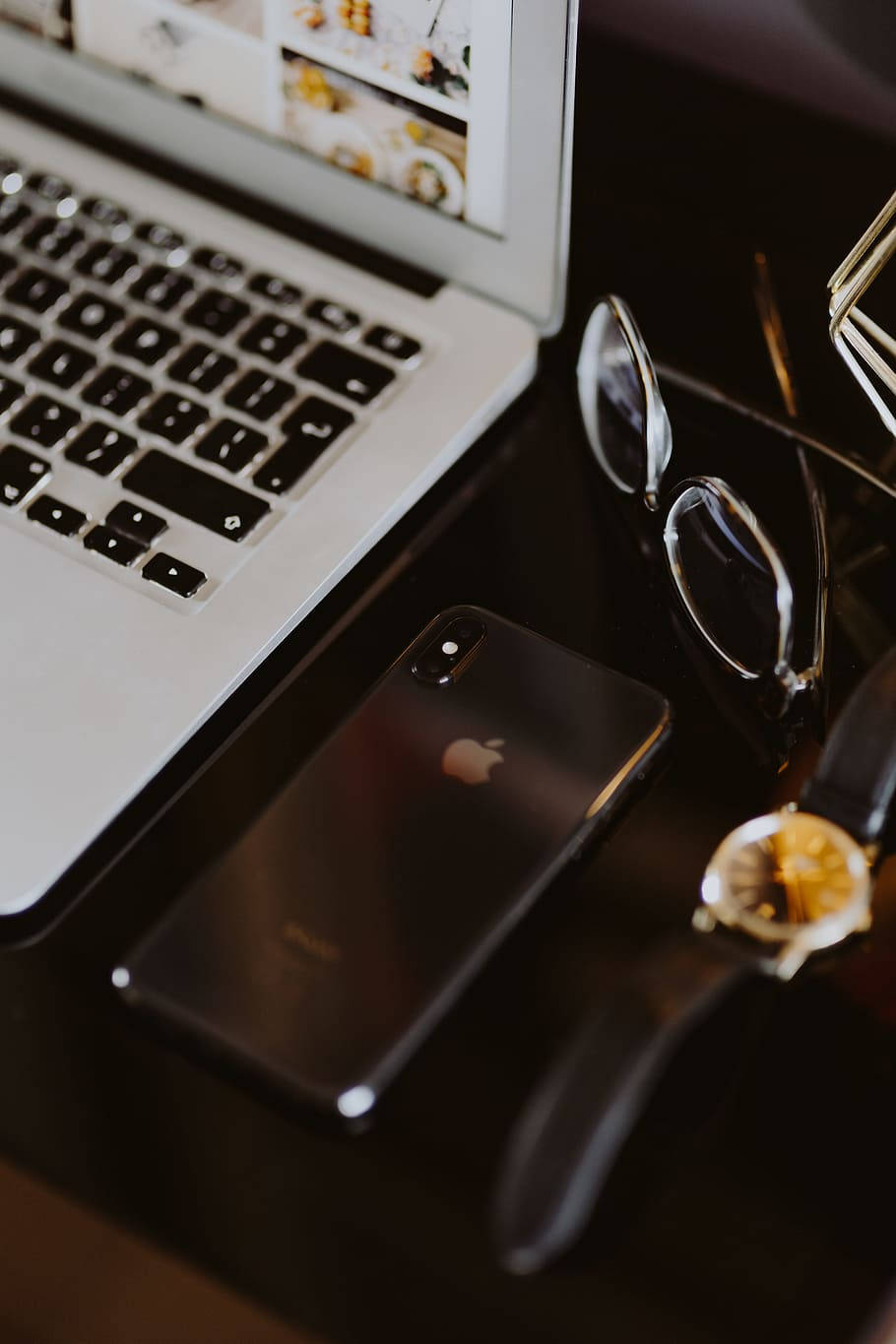 美學 桌布 壁紙 — 一部時尚的銀色筆記型電腦、黑色iPhone和金色手錶放在深色木桌上，旁邊有閱讀眼鏡。
