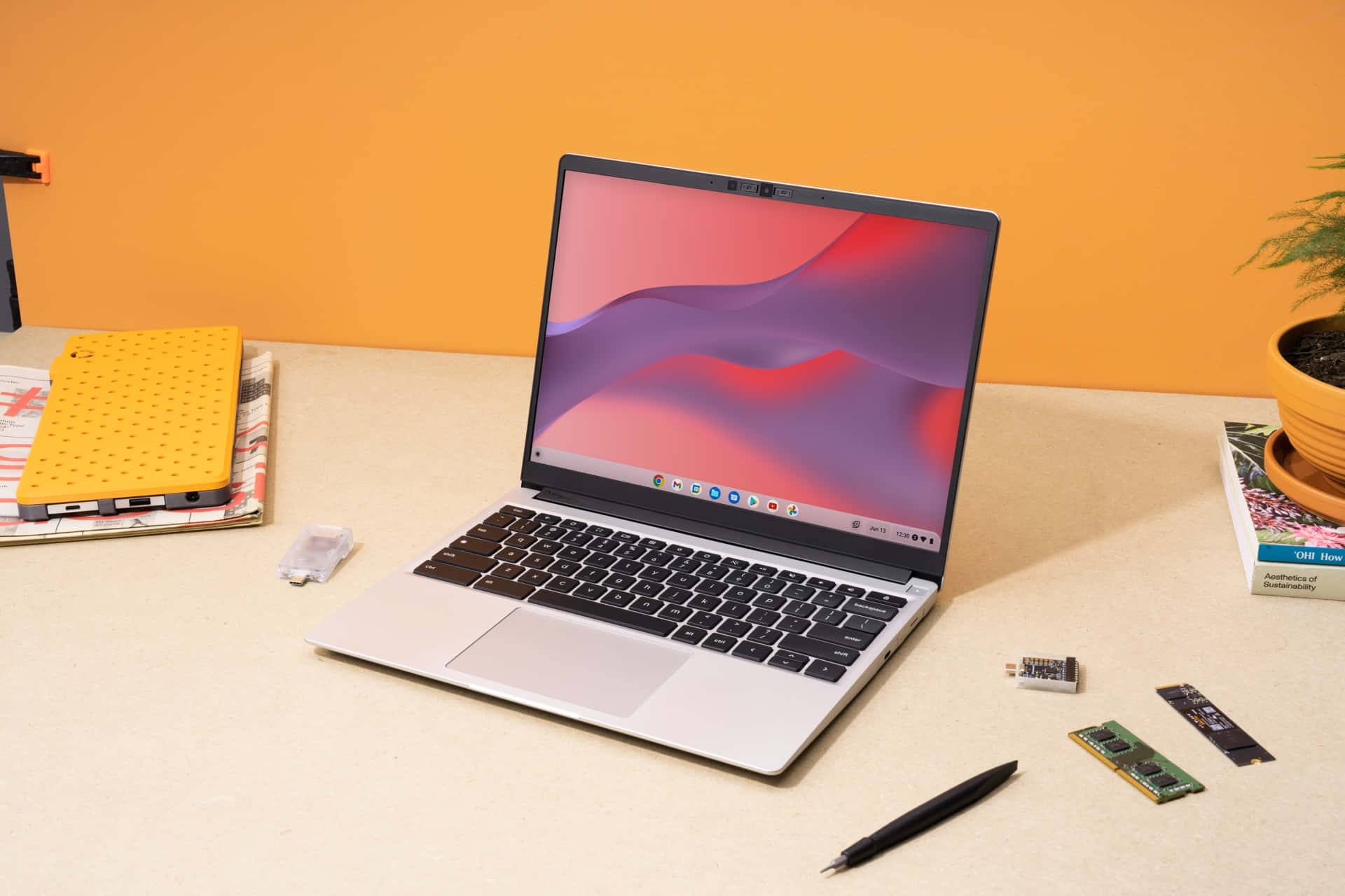 Chromebook 桌布 壁紙 — 一部時尚的銀色筆記型電腦在整潔的桌子上顯示充滿活力的抽象桌布，旁邊是科技配件和盆栽植物。