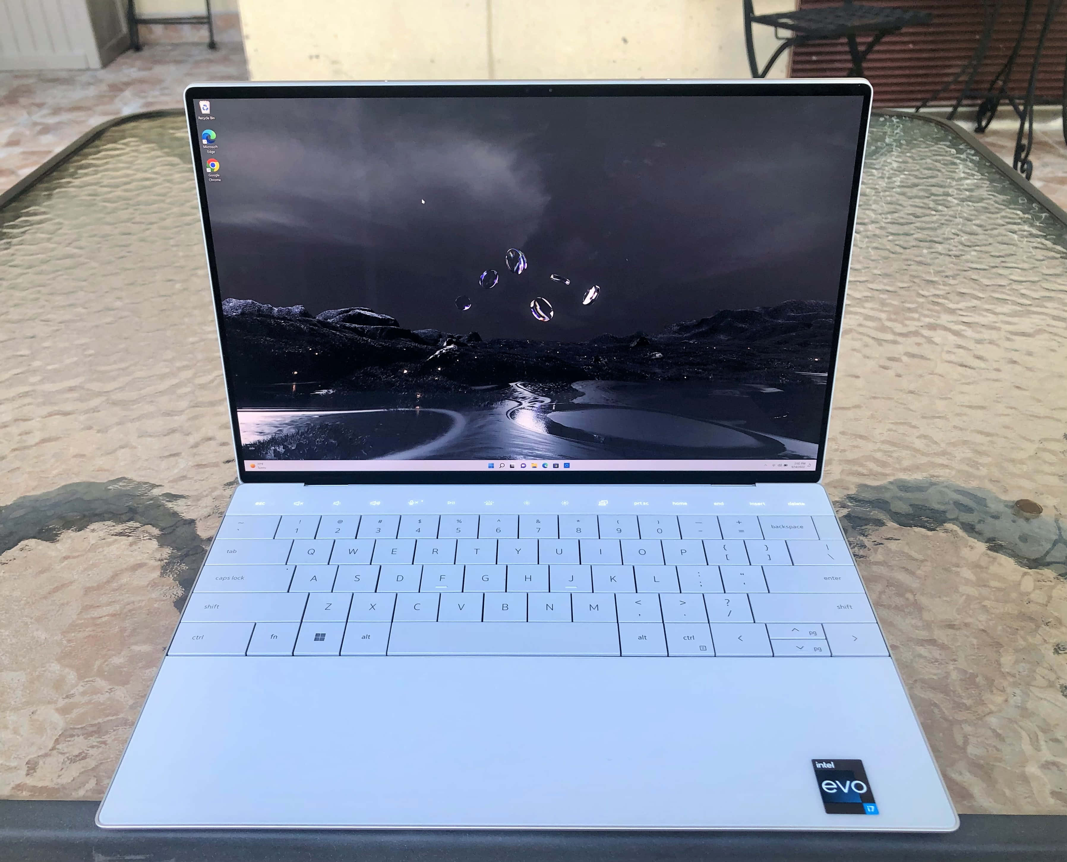 Dell 桌布 壁紙 — 一部時尚的銀色筆記型電腦打開在紋理玻璃桌上，顯示黑暗的山脈桌布，帶有發光球體和反射路徑。