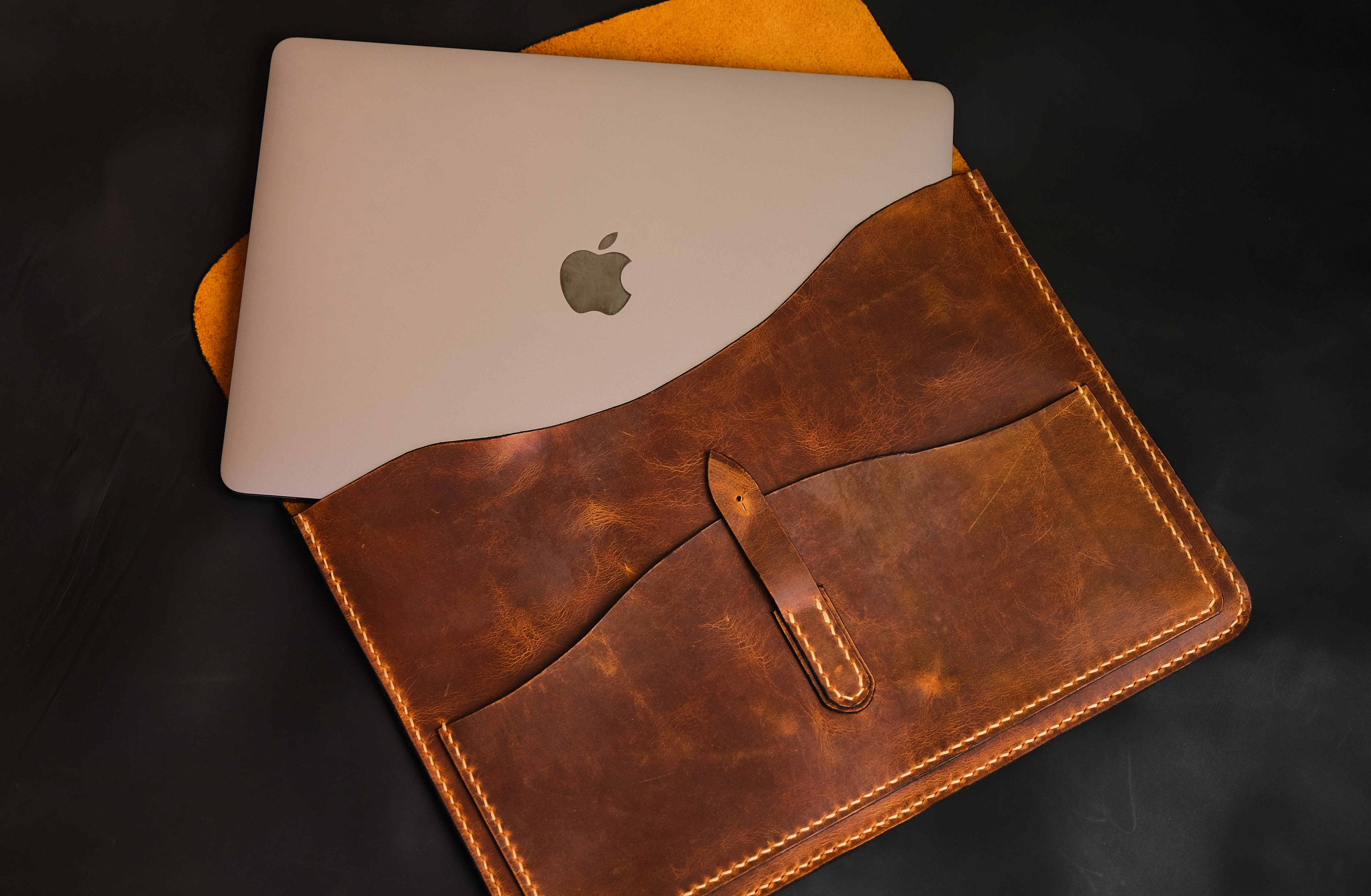 MacBook 動態圖片 桌布 壁紙 — 一部時尚的銀色筆記型電腦放在一個帶有可見縫線和翻蓋扣的濃郁棕色皮革保護套中，背景為深色。