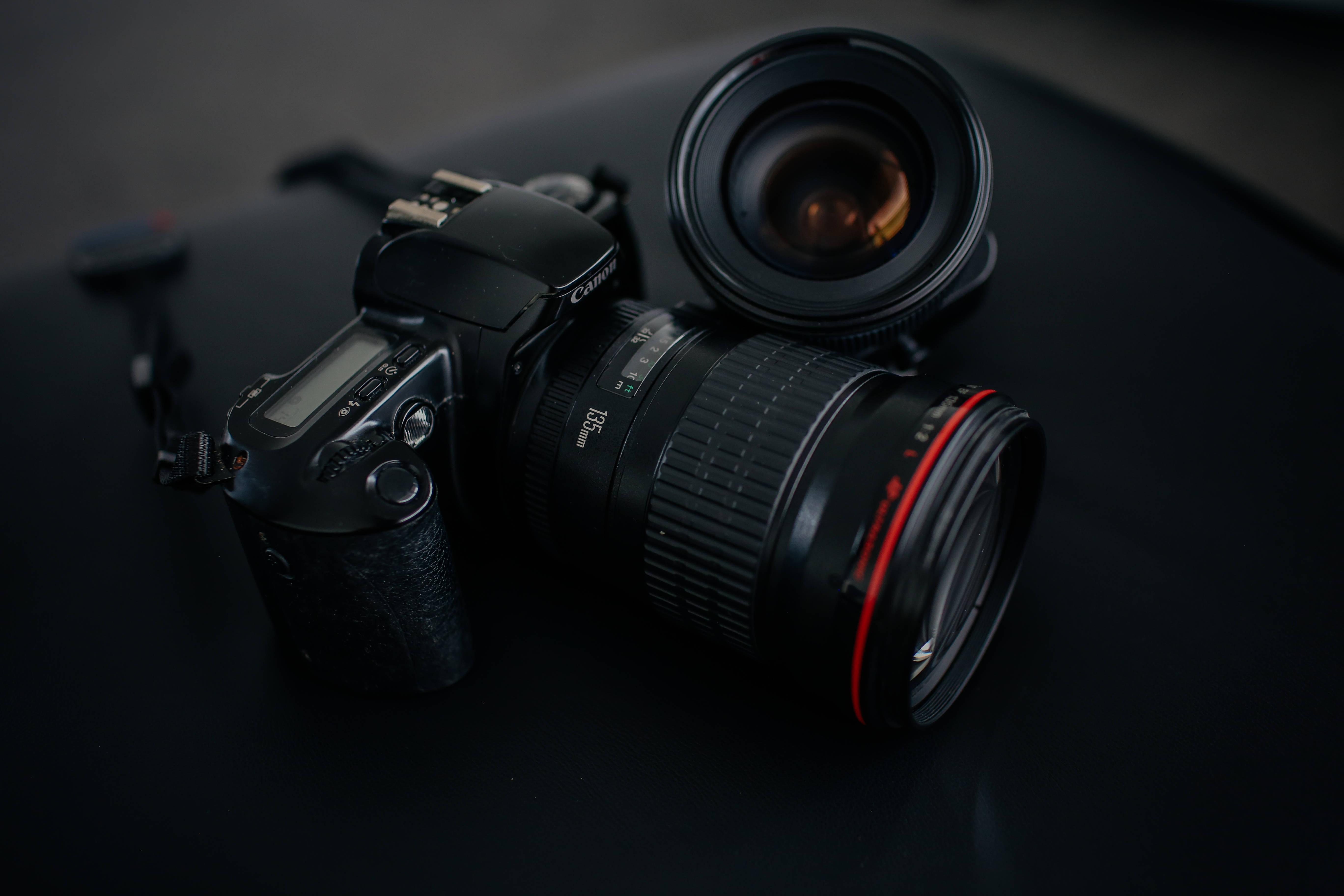 相機 桌布 壁紙 — 一部時尚的黑色 DSLR 相機，帶有紅色環形鏡頭，在昏暗的燈光下，旁邊放著一個分離的鏡頭，放在深色表面上。