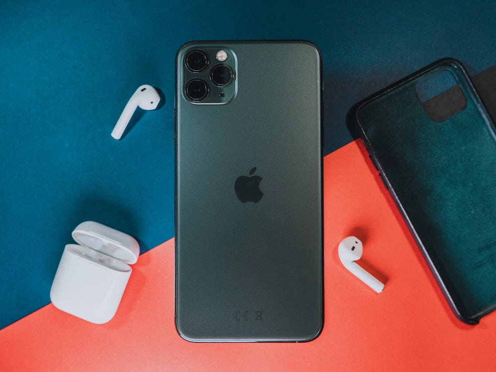 iPhone 11 桌布 壁紙 — 一部時尚的黑色 iPhone，搭配白色 AirPods 和保護殼，排列在鮮明的青綠色和珊瑚色背景上。