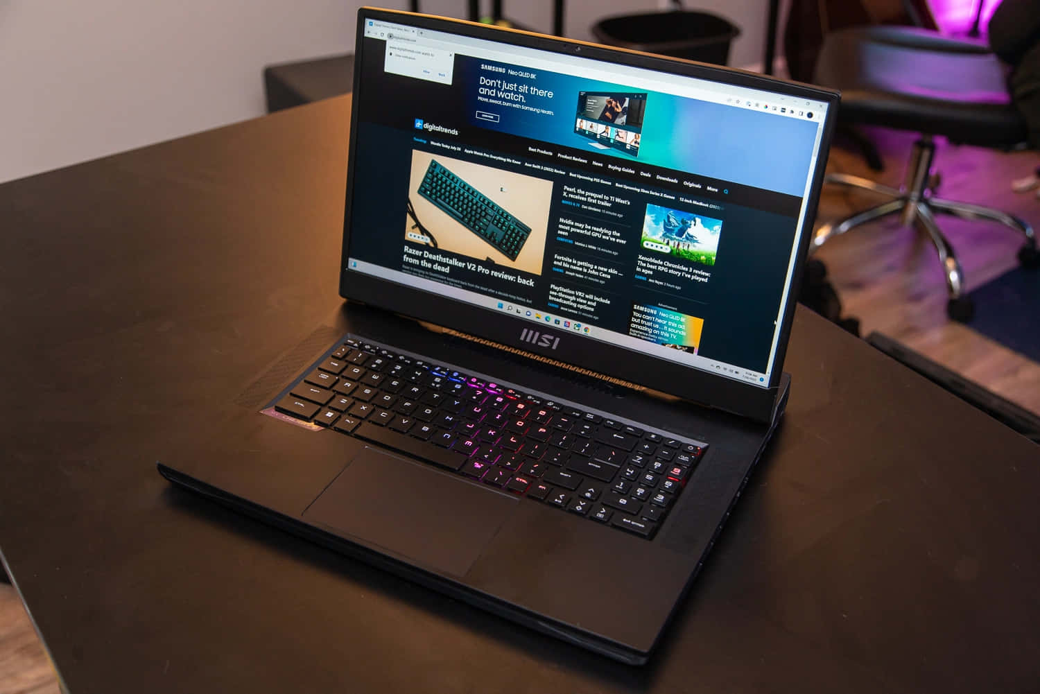 微星 桌布 壁紙 — 一部時尚的黑色 MSI 遊戲筆記型電腦，配備 RGB 鍵盤，打開放在深色木桌上，顯示科技評論網站。