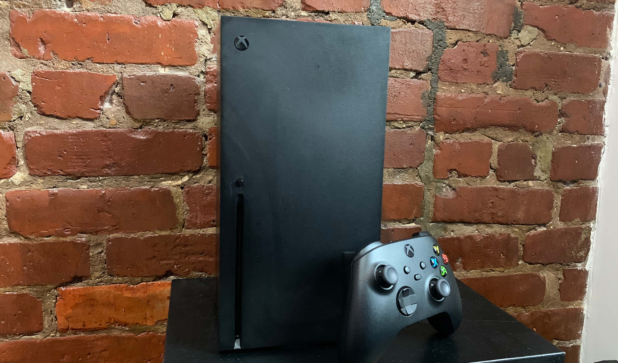 Xbox 桌布 壁紙 — 一部時尚的黑色 Xbox Series X 主機和控制器放在黑色支架上，背景是質樸的紅磚牆。