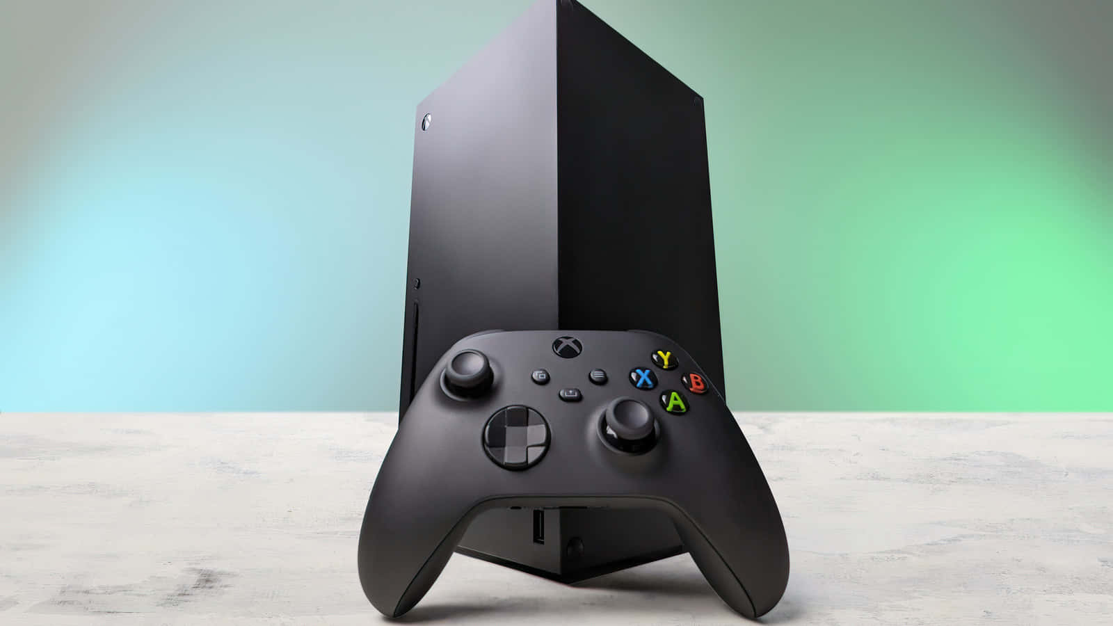Xbox 桌布 壁紙 — 一部時尚的黑色 Xbox Series X 主機直立放置，旁邊是其相配的控制器，置於淺色木質表面上，背景為柔和的藍綠漸變色。