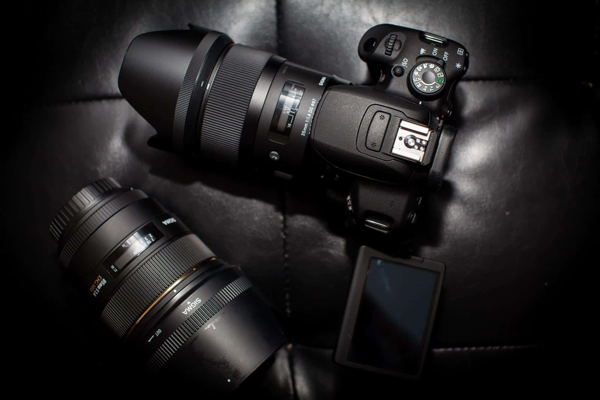 Dslr 桌布 壁紙 — 一部時尚的黑色DSLR相機配備兩個鏡頭，在戲劇性的低調燈光下放置在深色皮革表面上。