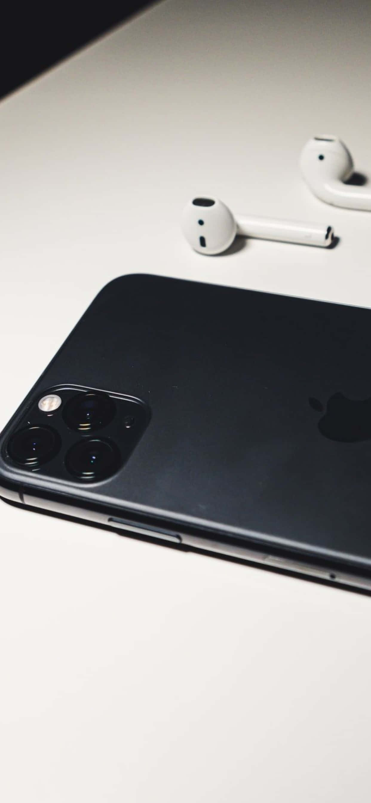 iPhone 11 Pro Max 桌布 壁紙 — 一部時尚的黑色iPhone，配備三鏡頭陣列，放在白色無線耳機旁邊，置於簡約的白色表面上。