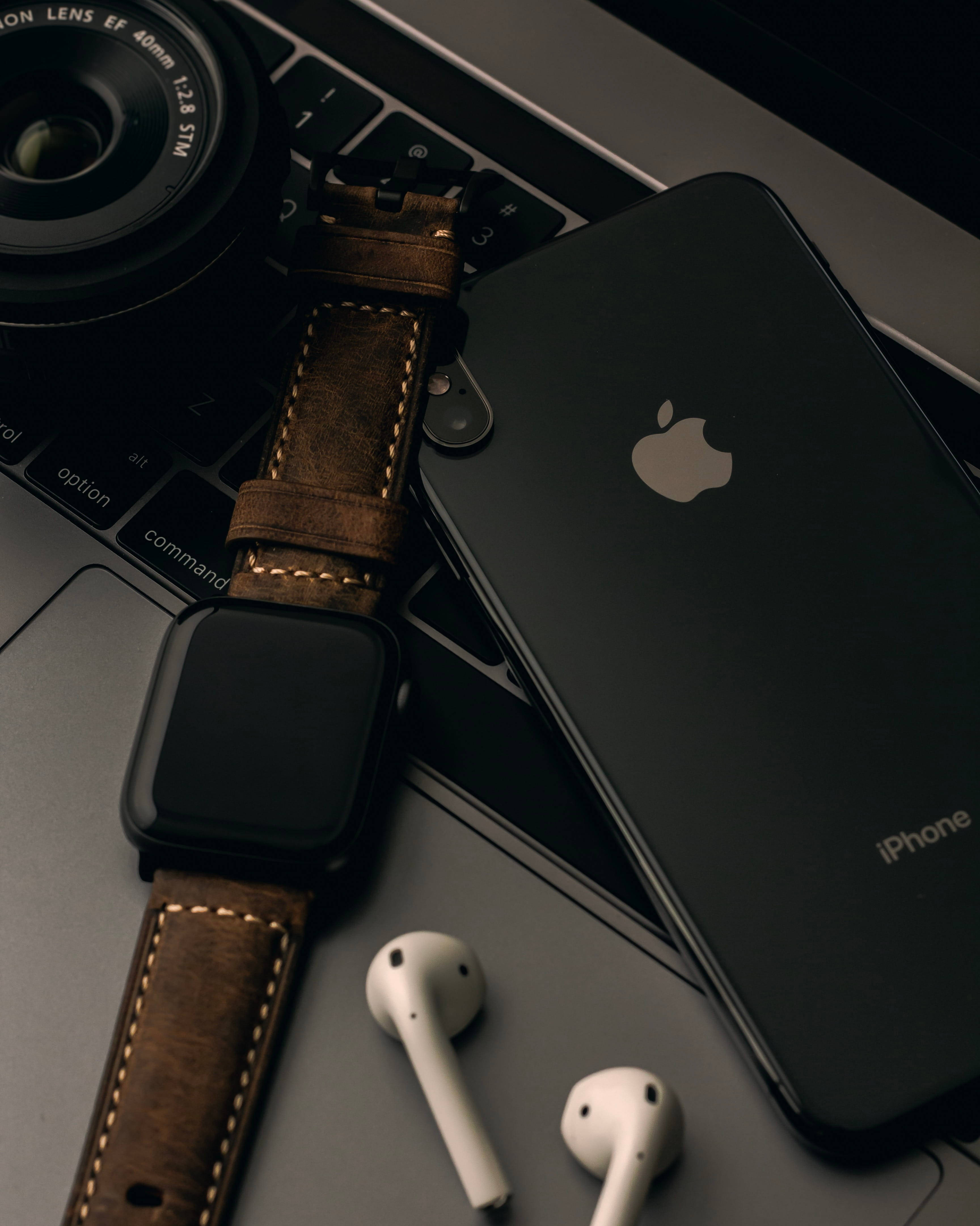 4K iPhone 11 桌布 壁紙 — 一部時尚的黑色iPhone和棕色皮革錶帶的智慧手錶放在筆記型電腦鍵盤上，旁邊有白色耳機和相機鏡頭。