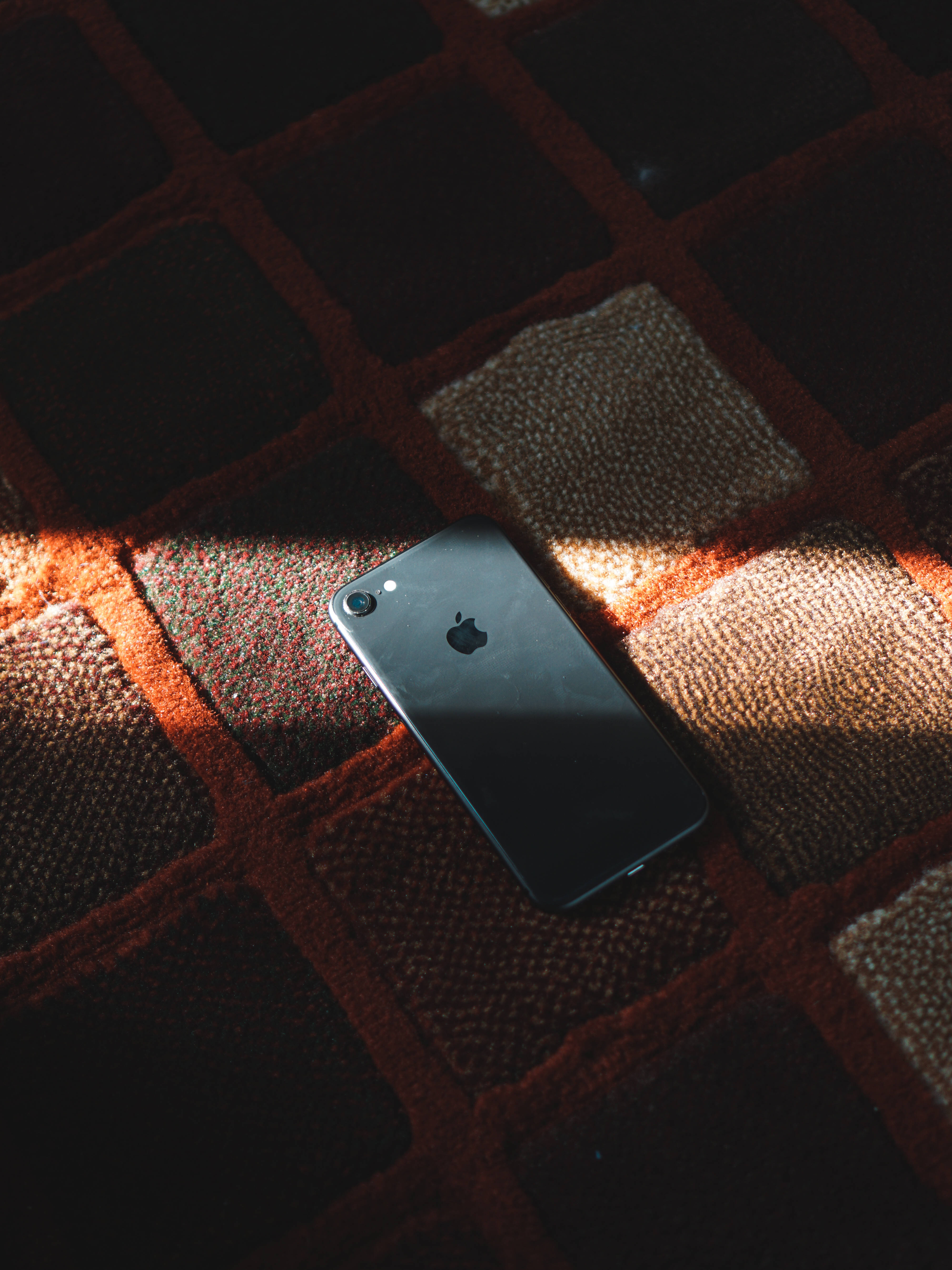 iPhone 8 桌布 壁紙 — 一部時尚的黑色iPhone放在一張質感豐富的紅米色格紋地毯上，沐浴在溫暖的陽光中。