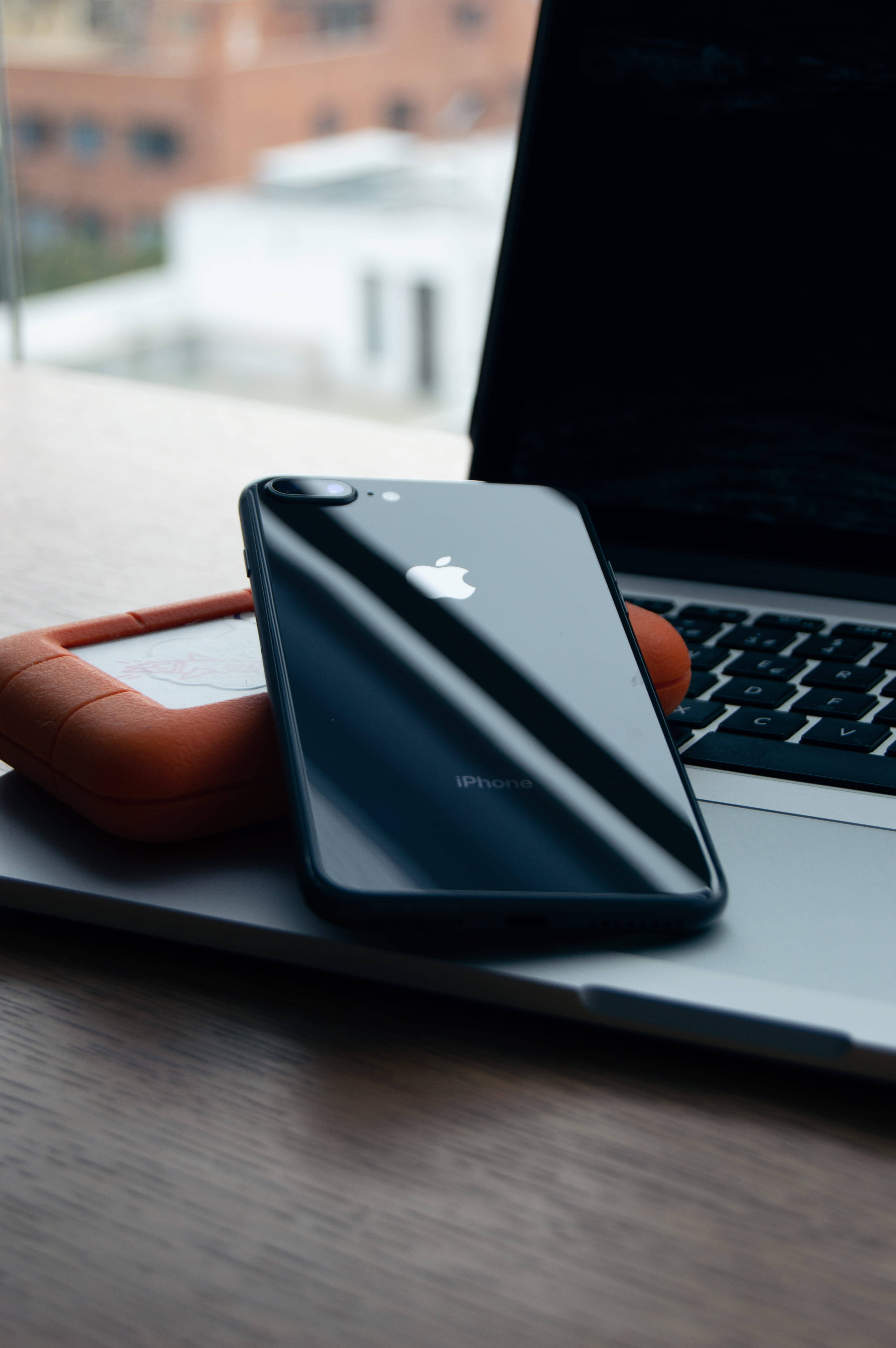 MacBook 動態圖片 桌布 壁紙 — 一部時尚的黑色iPhone放在銀色筆記型電腦旁，旁邊有一個橙色手機殼，置於木桌上，背景是模糊的城市建築。