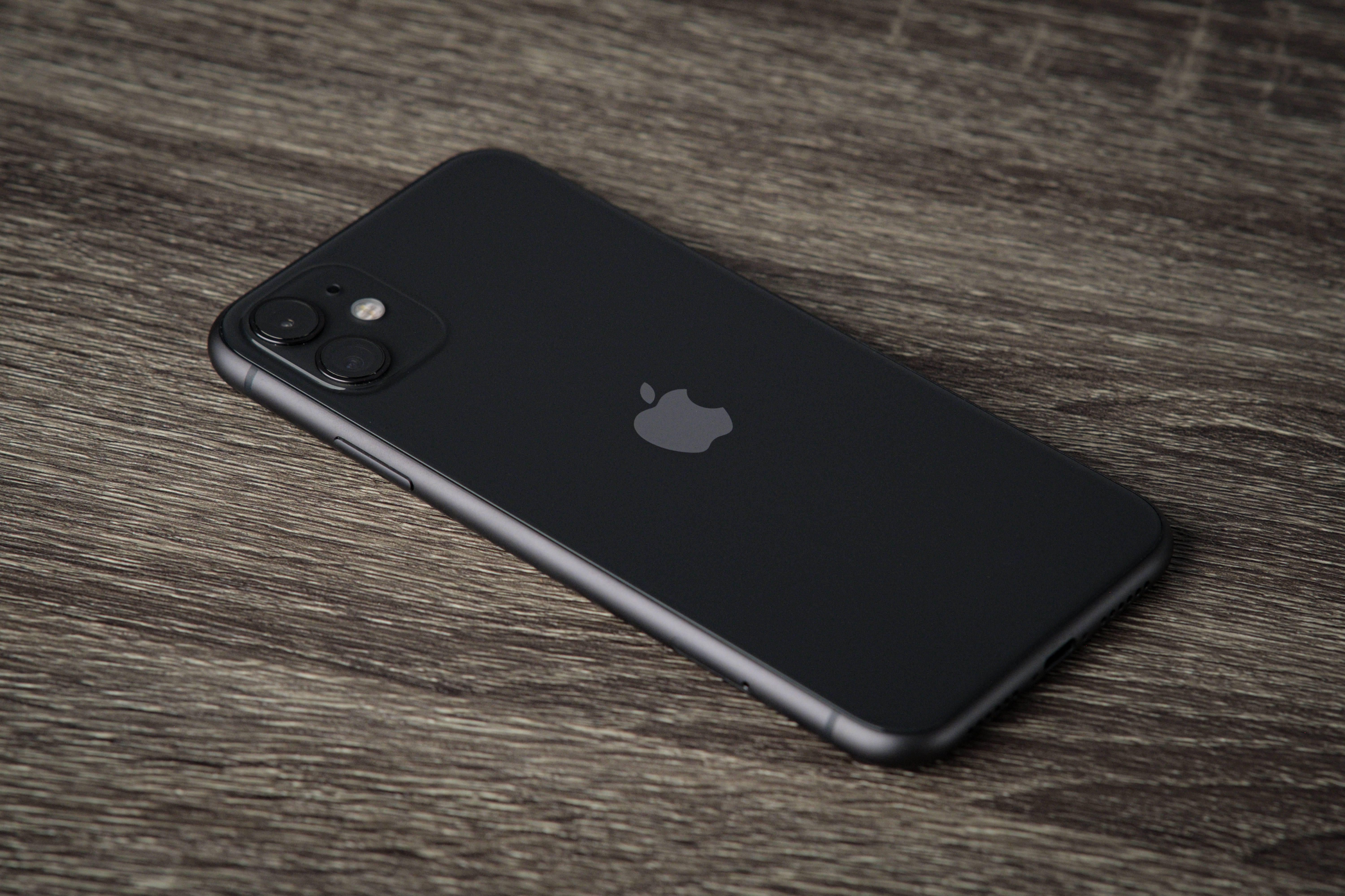 iPhone 11 Black 桌布 壁紙 — 一部時尚的黑色iPhone斜放在有紋理的灰色木質表面上，柔和自然光線照射。