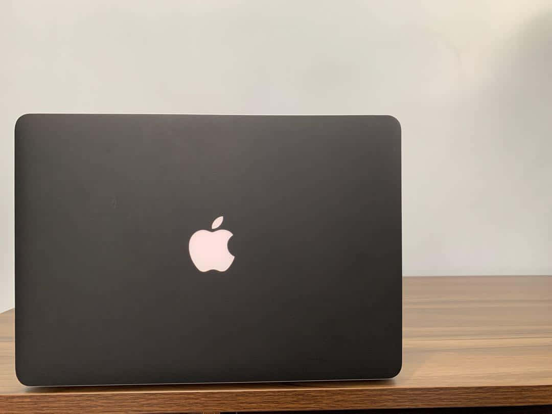 Black Macbook 桌布 壁紙 — 一部時尚的黑色MacBook，帶有發光的蘋果標誌，闔上放在木桌上，背景是純白牆壁。