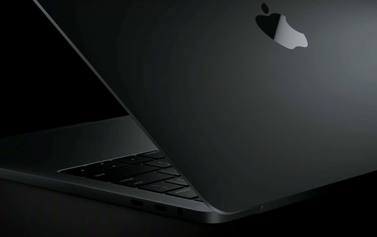 Black Macbook 桌布 壁紙 — 一部時尚的黑色MacBook筆記型電腦在陰影中傾斜，其銀色Apple標誌在深色背景下柔和發光。