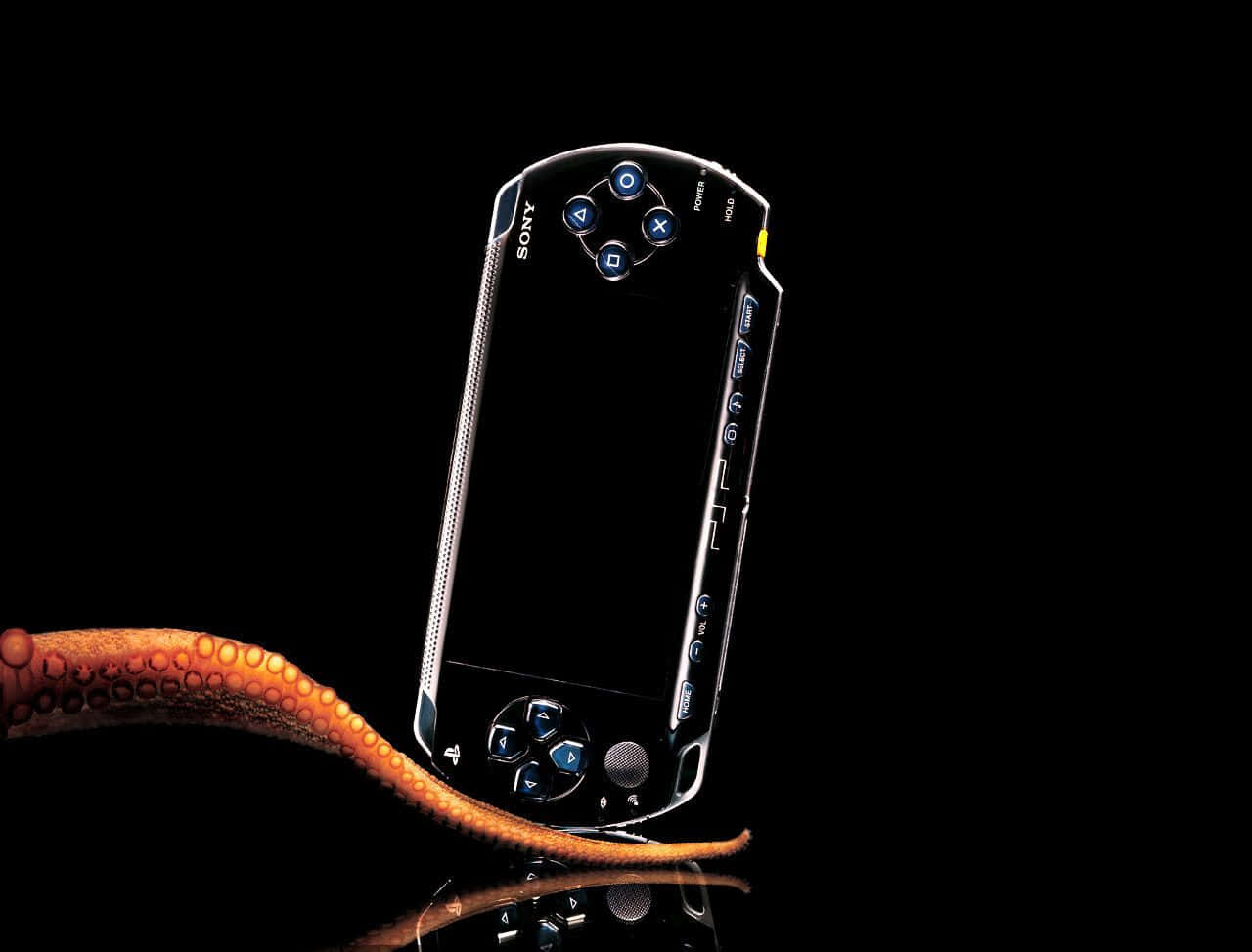 Psp 桌布 壁紙 — 一部時尚的黑色Sony PSP放在反光表面上，旁邊有一條橙色章魚觸手在黑暗背景中蜷曲。