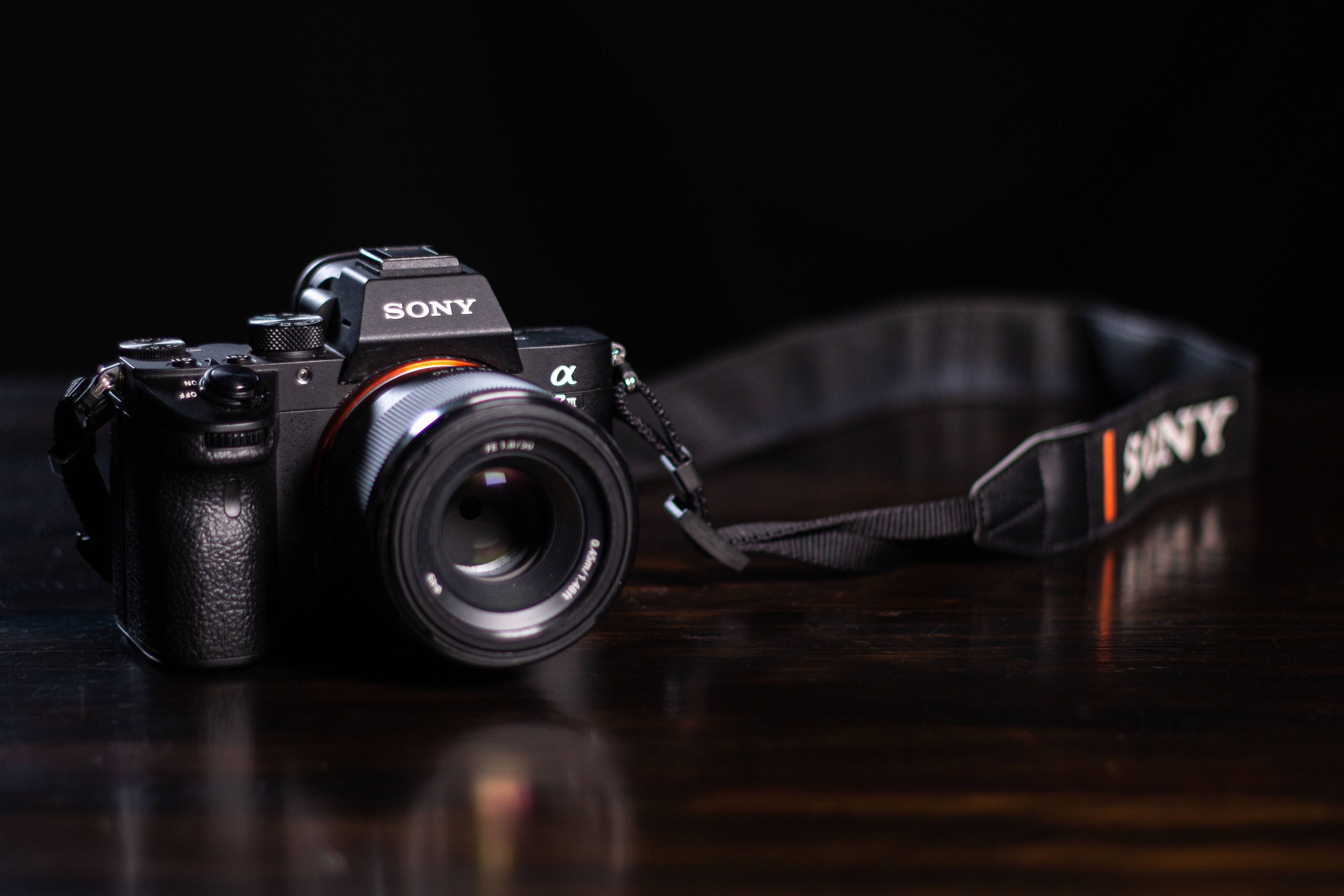 Simple HD 桌布 壁紙 — 一部時尚的黑色Sony相機連鏡頭，放置在黑色背景前的深色木質表面上。