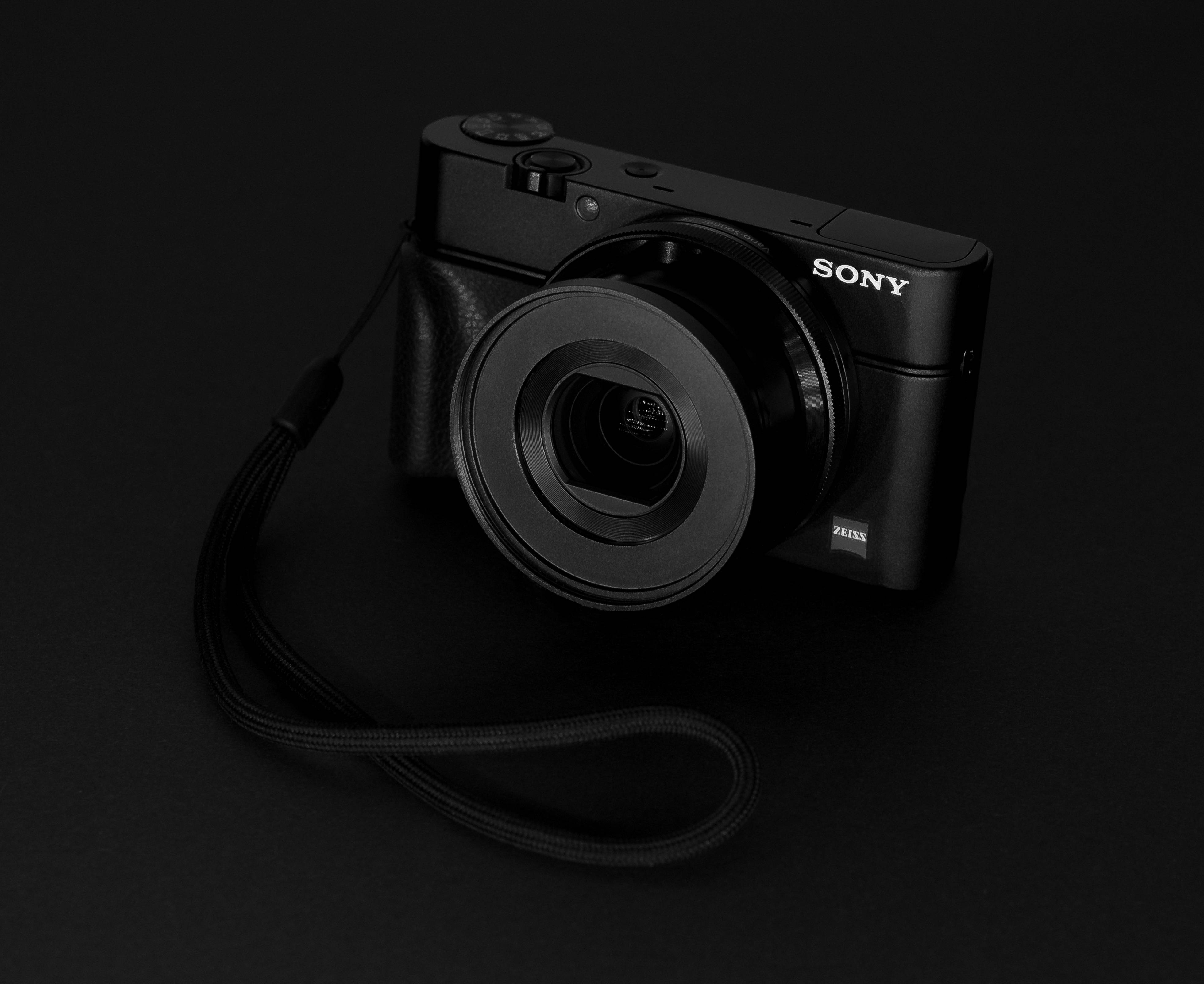 Sony 桌布 壁紙 — 一部時尚的黑色Sony相機配備Zeiss鏡頭，放置在深色表面上，其背帶纏繞在一旁，在戲劇性的工作室燈光下。