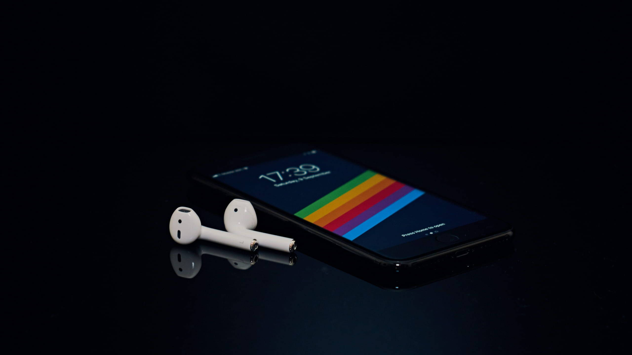 AirPods 桌布 壁紙 — 一部時尚的黑色智慧型手機，帶有彩虹條紋桌布和白色耳塞，放置在反光的深色表面上。