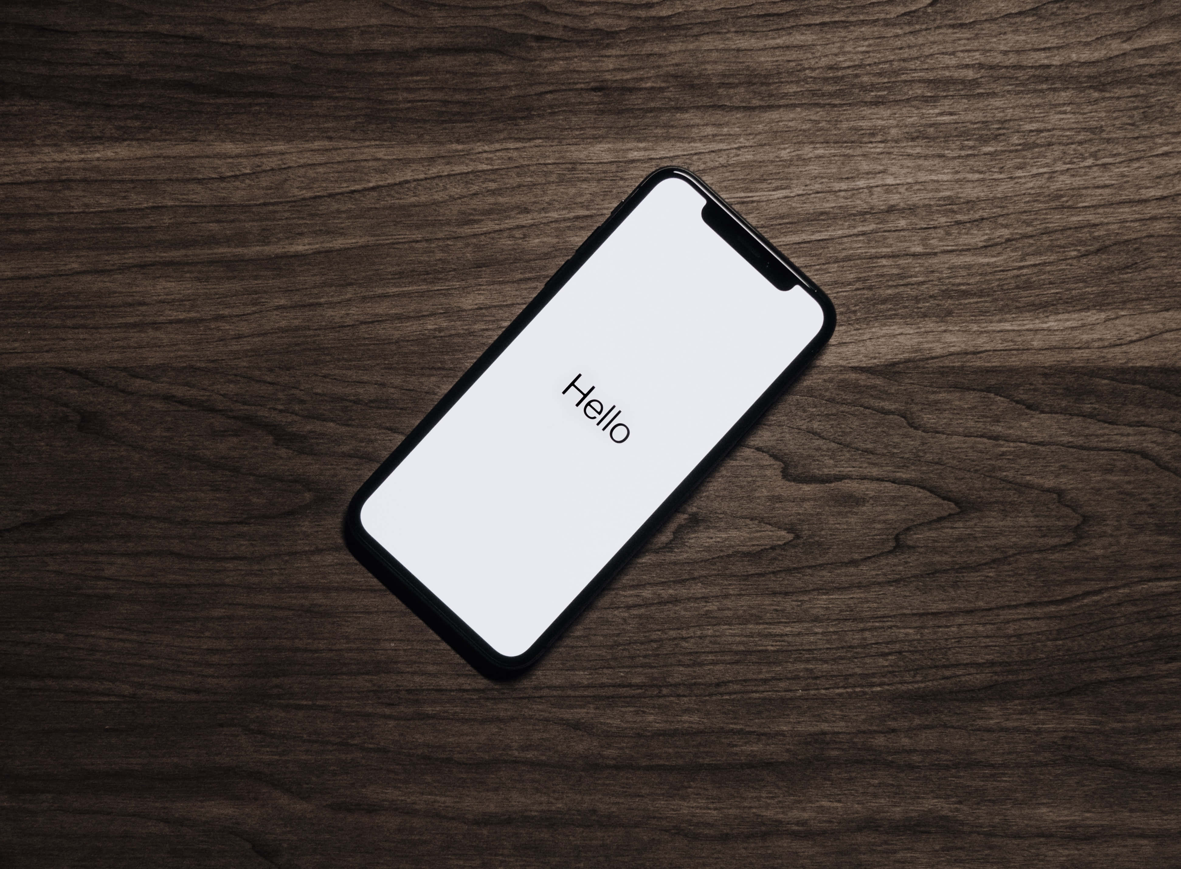 Device 桌布 壁紙 — 一部時尚的黑色智慧型手機，白色螢幕上顯示「hello」一詞，對角放置在深色木紋表面上。