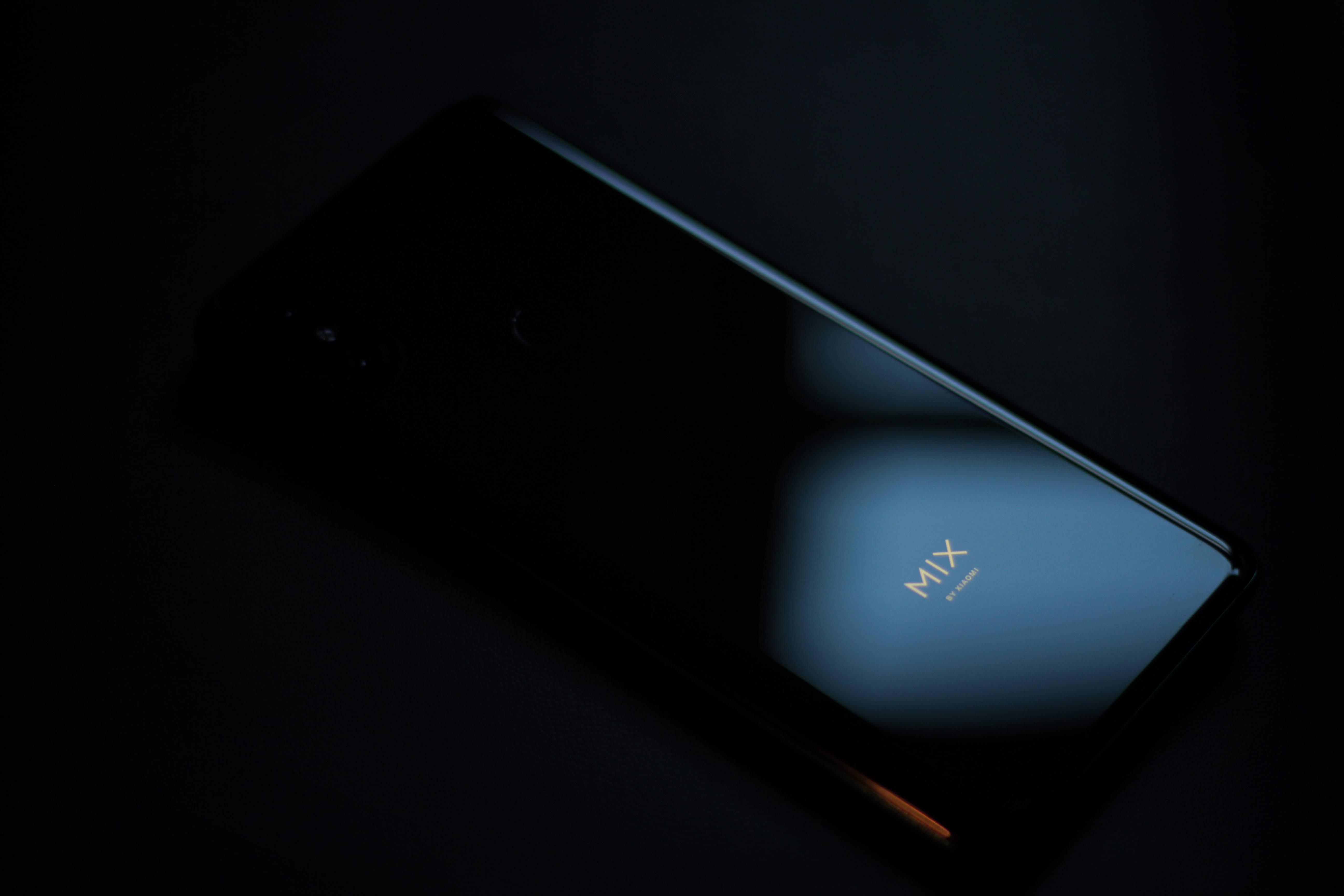 Xiaomi 桌布 壁紙 — 一部時尚的黑色智慧型手機在近乎黑暗中放置，藍色發光反射在其彎曲螢幕上，揭示金色的MIX字樣。