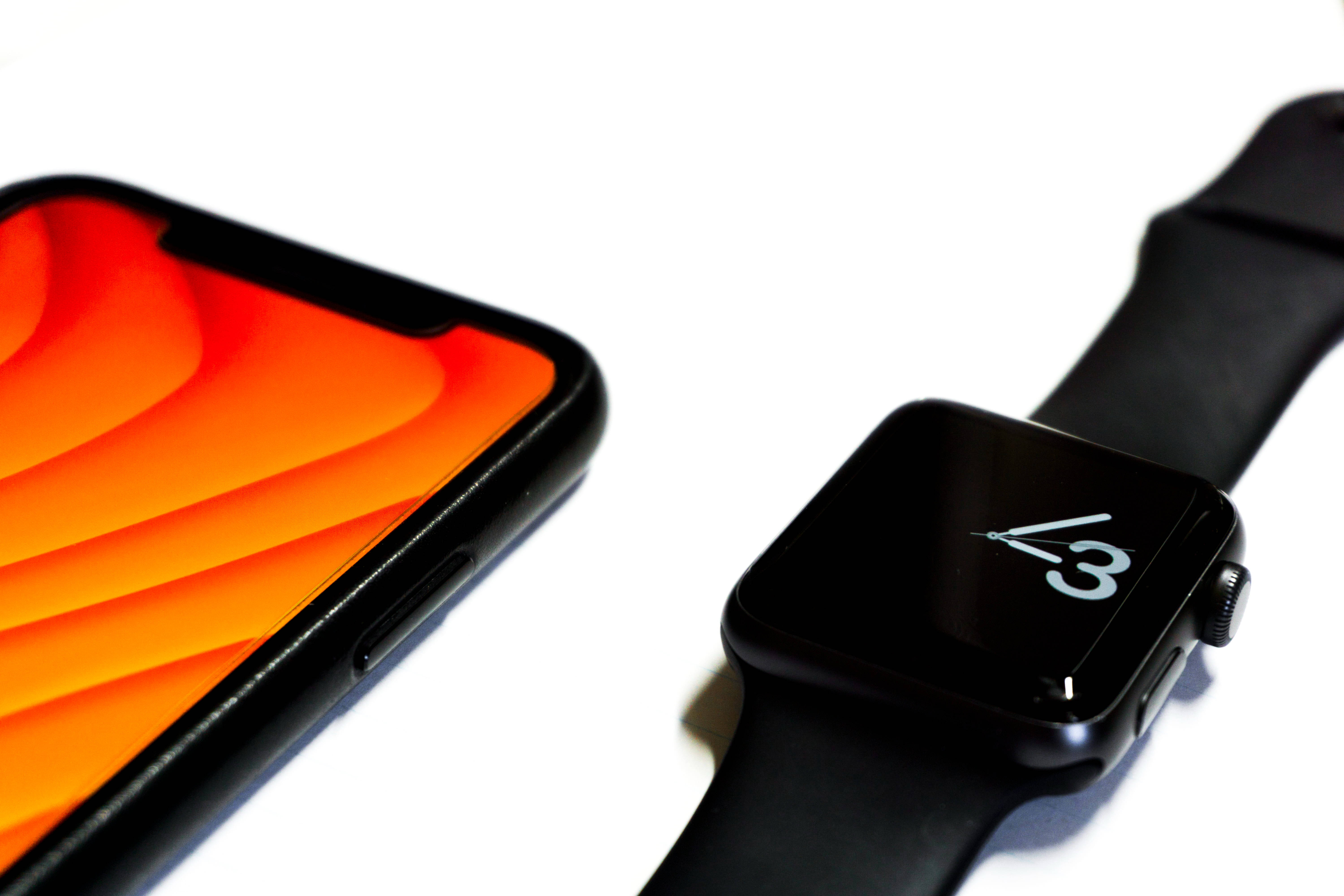 Apple Watch 動態 桌布 壁紙 — 一部時尚的黑色智慧型手機帶有充滿活力的橙色漸變壁紙，旁邊放著一隻黑色Apple Watch，在簡約的白色表面上顯示時間4:30。