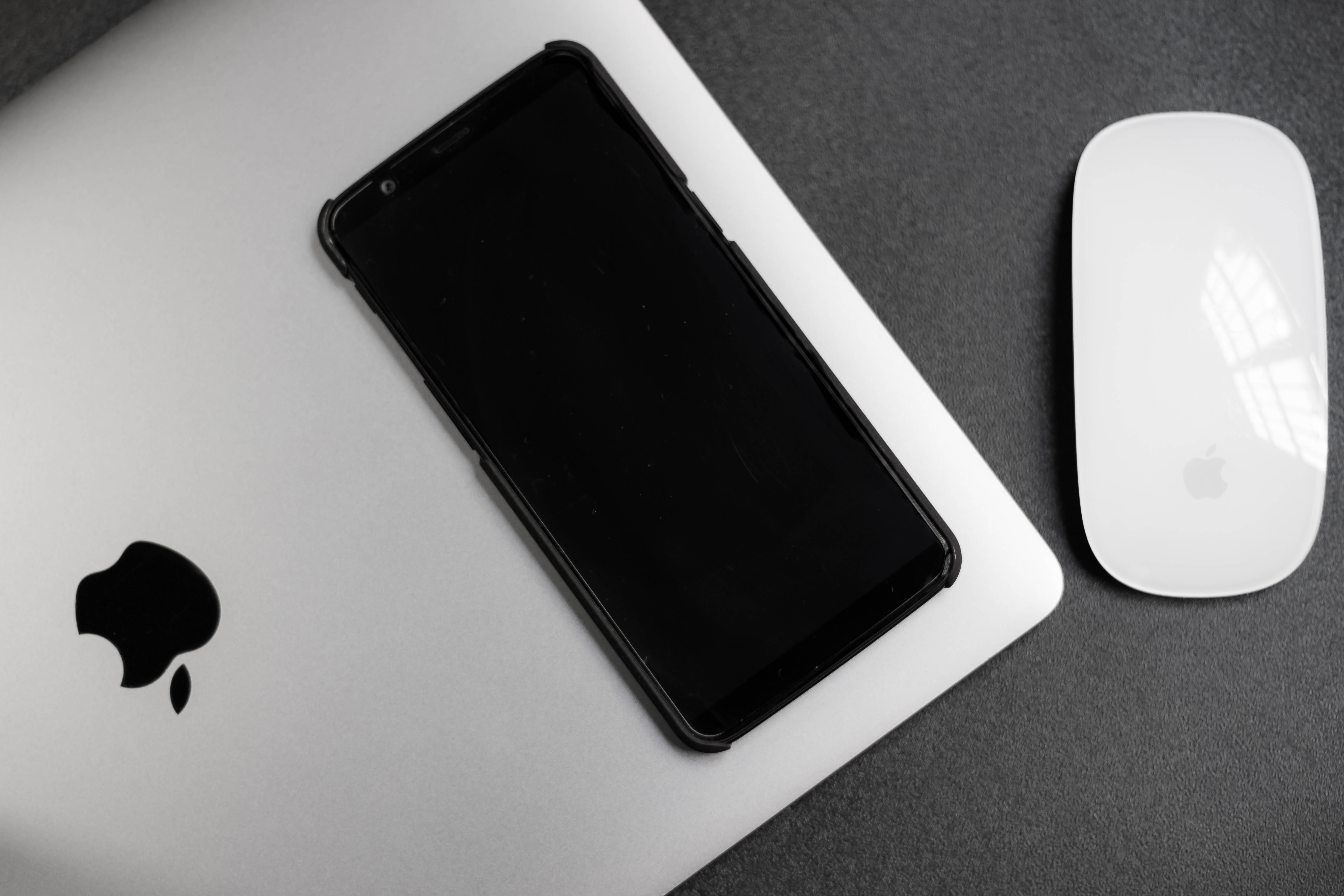 Black Android 桌布 壁紙 — 一部時尚的黑色智慧型手機放在銀色筆記型電腦上，旁邊有一個白色滑鼠，置於深灰色表面上。