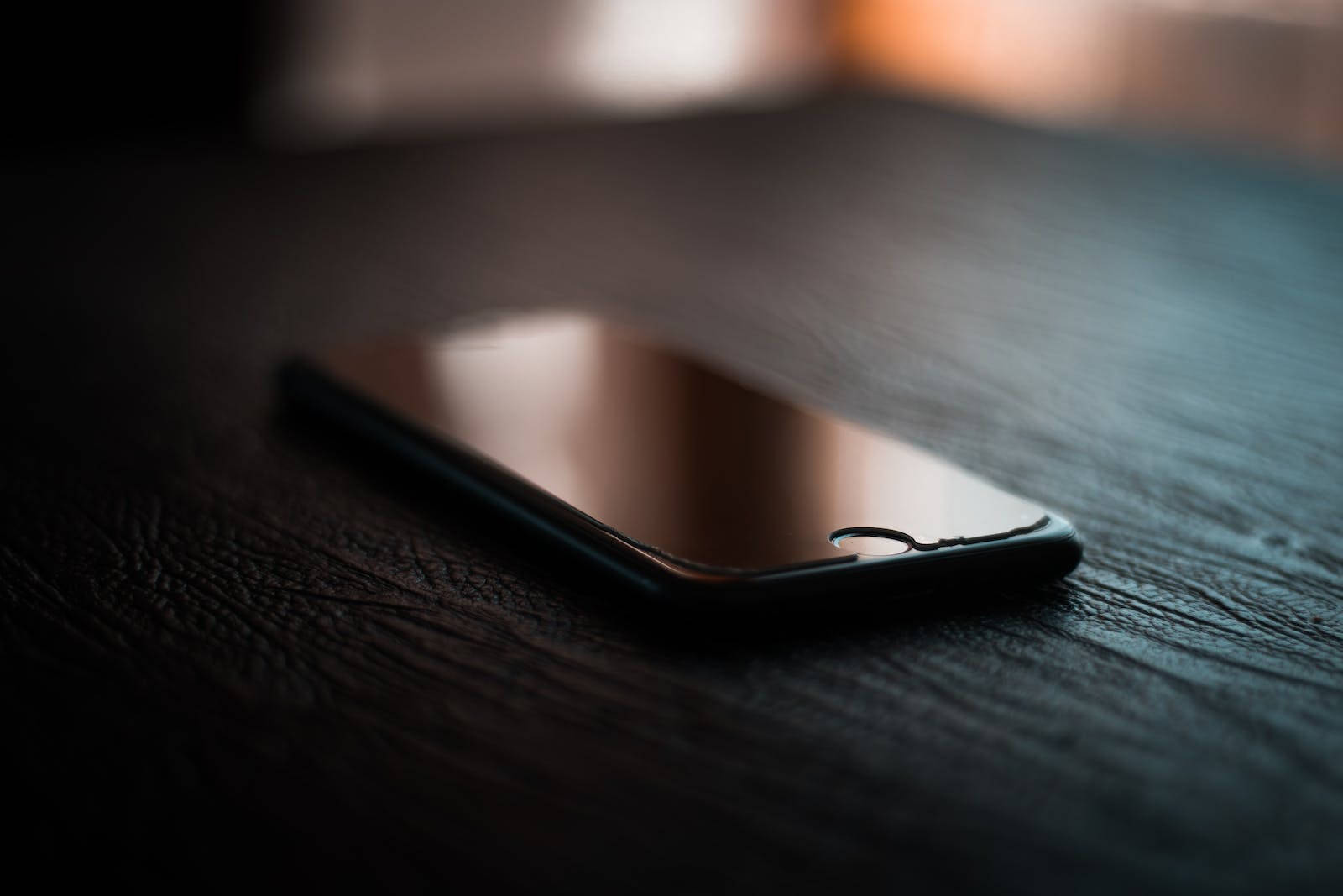 深色 iPhone 桌布 壁紙 — 一部時尚的黑色智慧型手機放置在深色紋理木質表面上，背景帶有柔和溫暖的燈光。