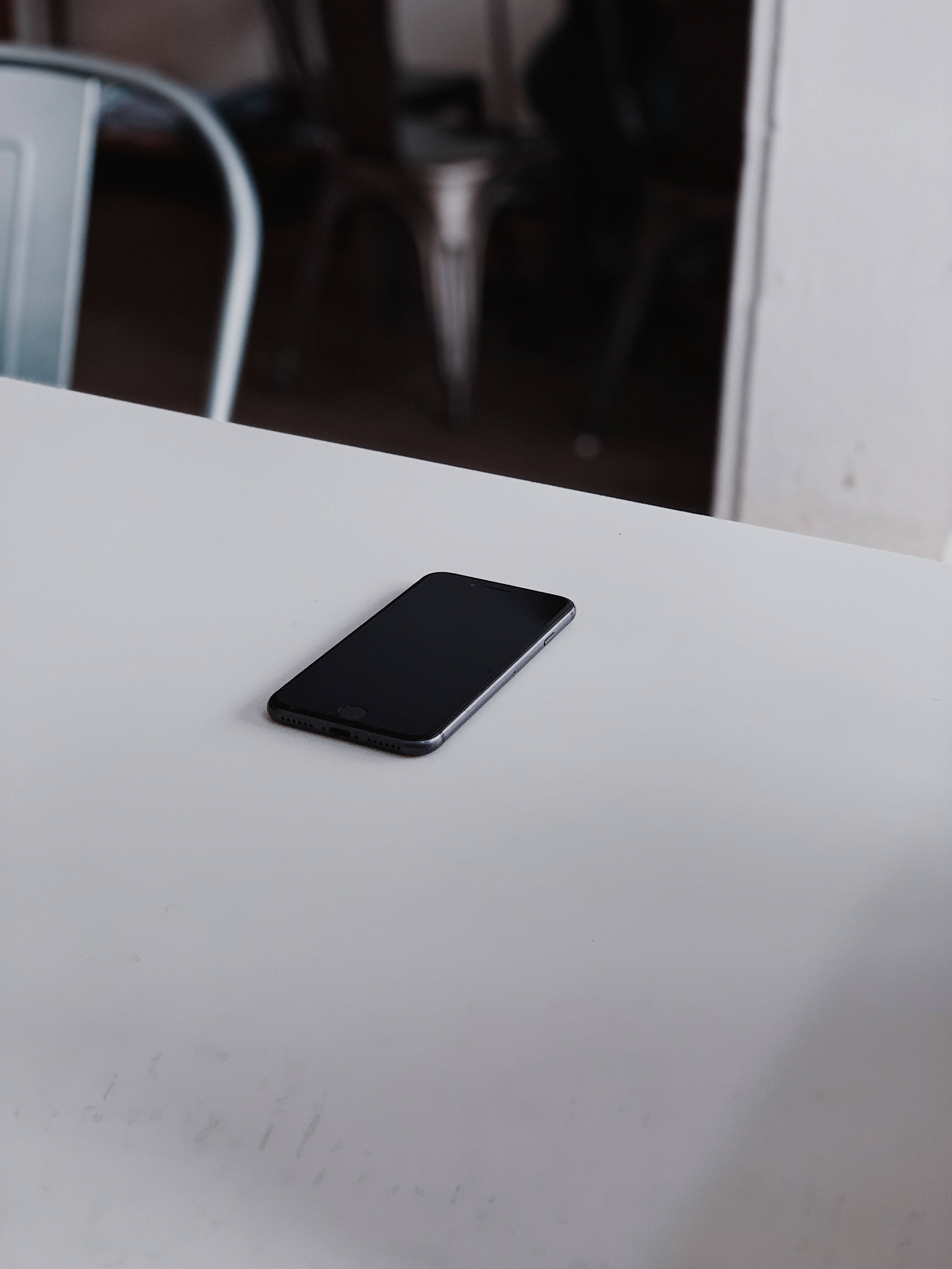 iPhone 8 桌布 壁紙 — 一部時尚的黑色智慧型手機獨自放在簡約的白色桌子上，背景有一張模糊的金屬椅子。