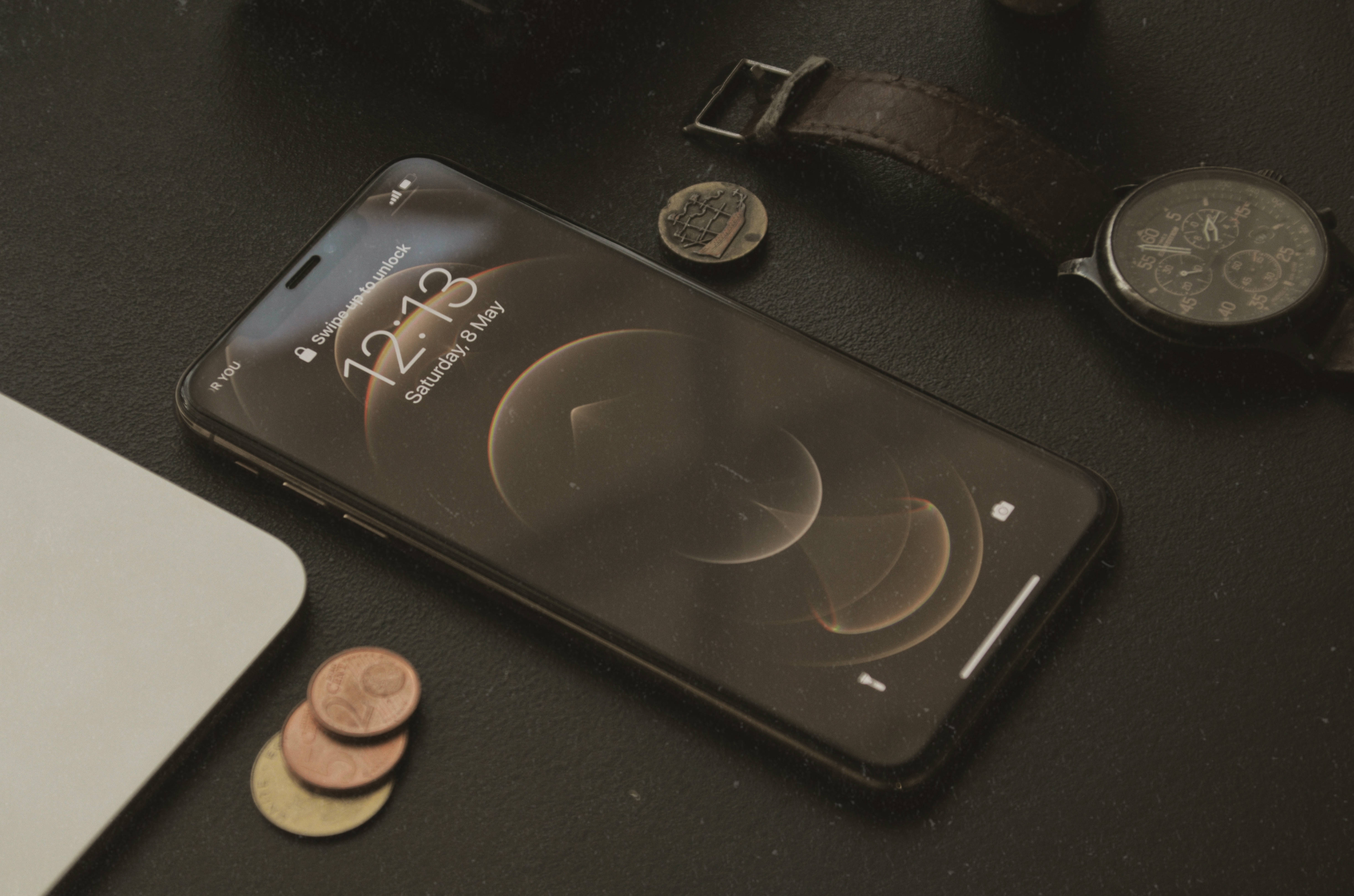 iPhone XS Max 桌布 壁紙 — 一部時尚的黑色智慧型手機顯示宇宙風格桌布，旁邊放著一隻復古手錶和散落的硬幣，置於深色紋理表面上。