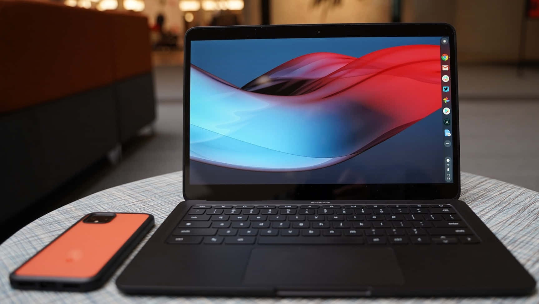 Chromebook 桌布 壁紙 — 一部時尚的黑色筆記型電腦，帶有充滿活力的紅色和藍色抽象桌布，放在圖案桌上，旁邊是一部橙色電話。