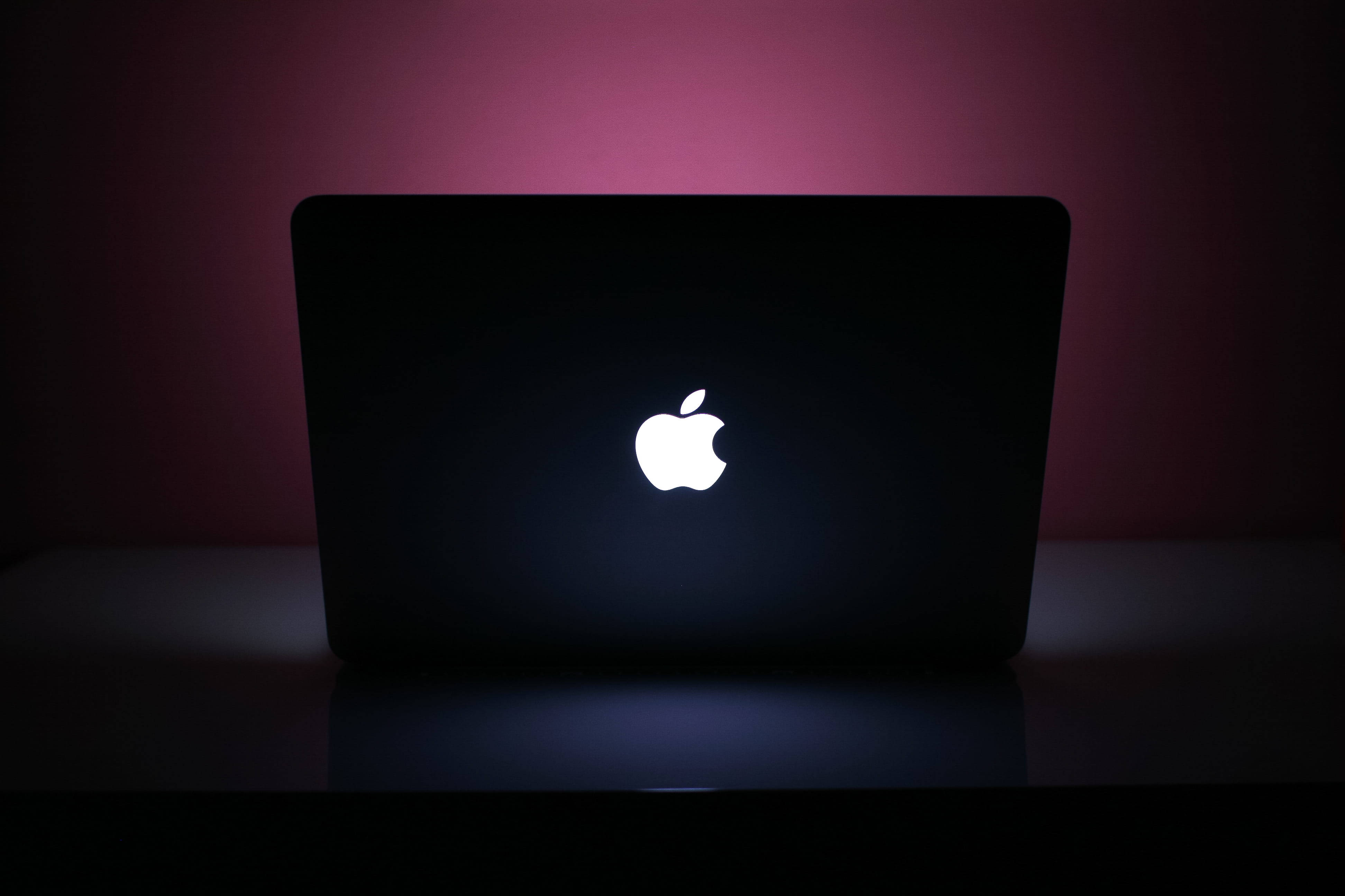 macbook pro 桌布 壁紙 — 一部時尚的黑色筆記型電腦，帶有發光的白色Apple標誌，放置在柔和粉紅色環境照明下的深色表面上。
