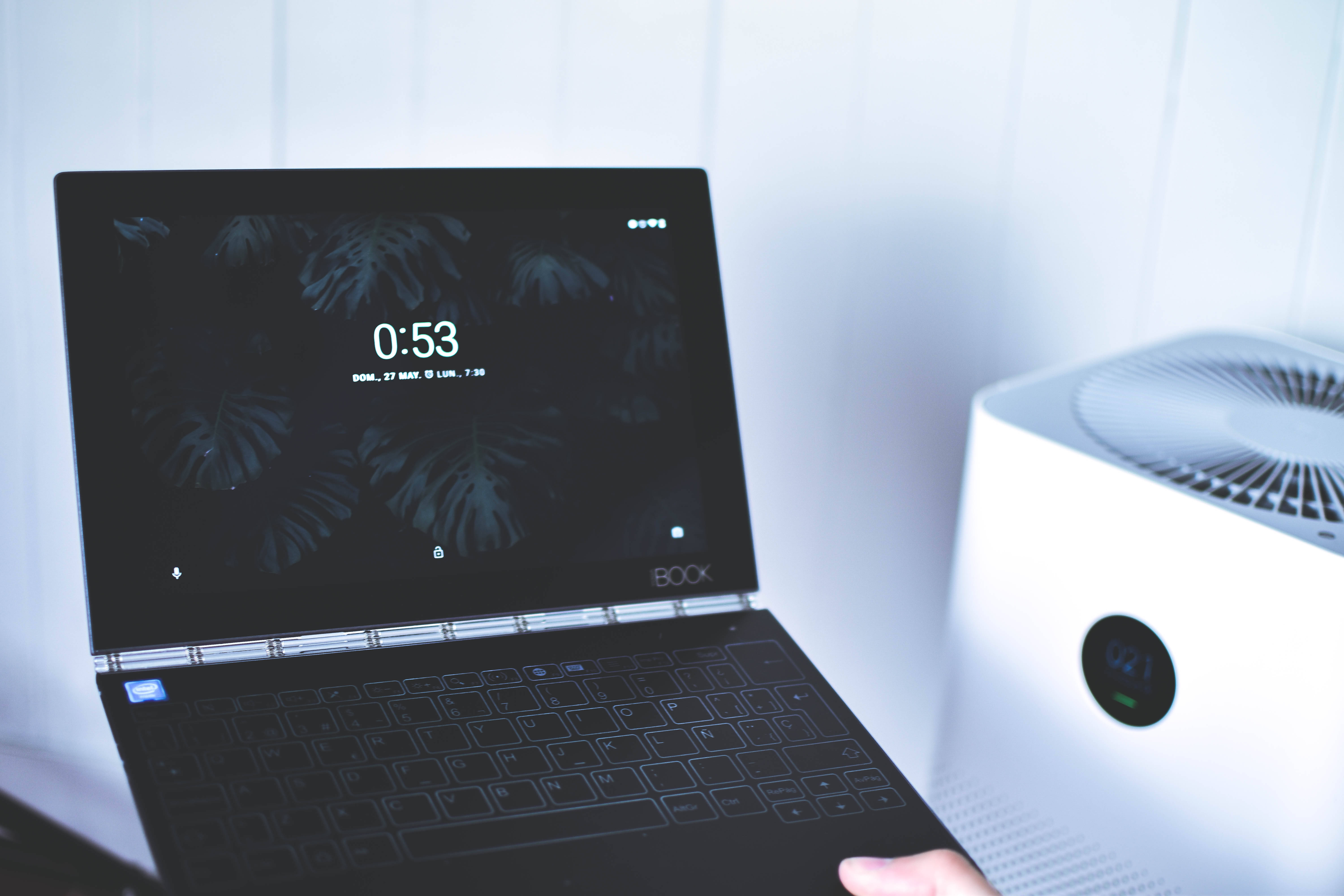 Xiaomi 桌布 壁紙 — 一部時尚的黑色筆記型電腦，帶有龜背竹葉壁紙和鎖定螢幕，旁邊放著一台白色空氣清淨機，位於明亮的簡約房間中。