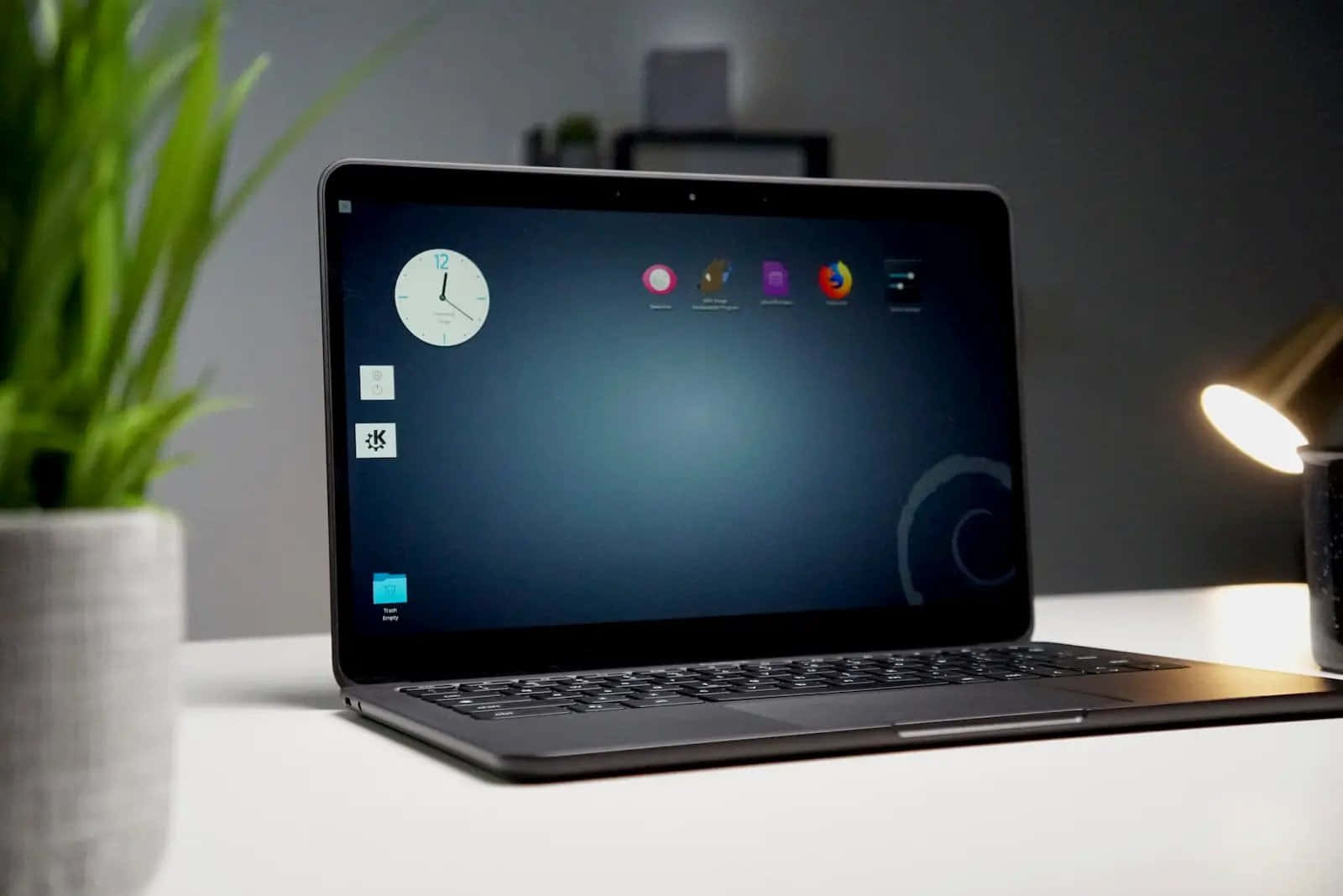 Chromebook 桌布 壁紙 — 一部時尚的黑色筆記型電腦打開放在白色桌子上，旁邊有一盆植物和一盞發光的檯燈，營造出簡約的工作空間。