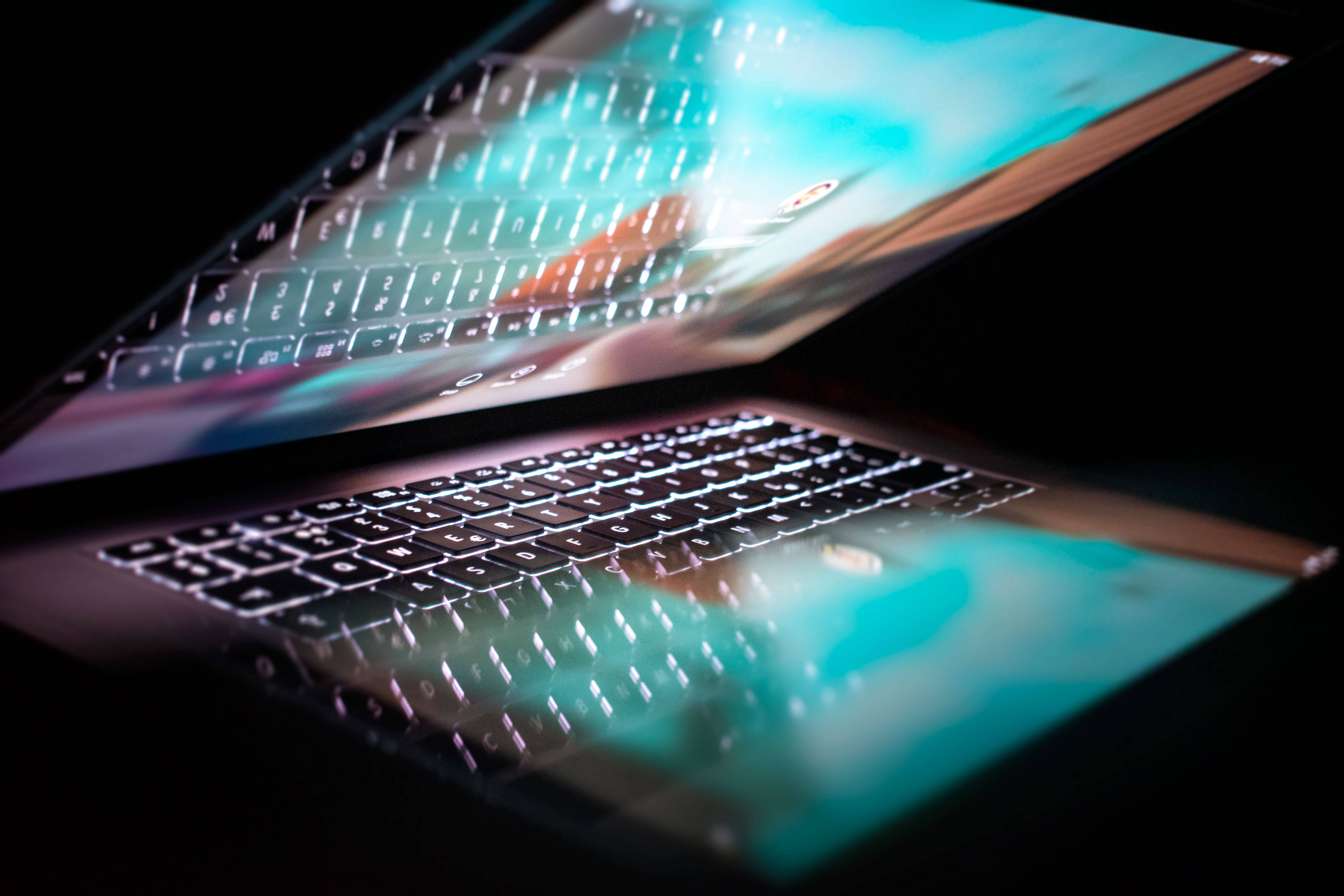 Computer Screen 桌布 壁紙 — 一部時尚筆記型電腦在黑暗中傾斜，帶有發光按鍵和柔和反射的鮮豔青綠色螢幕。