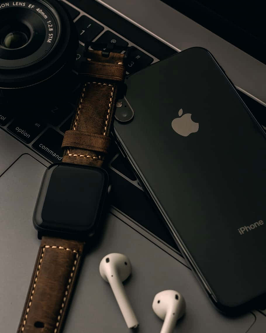 蘋果 桌布 壁紙 — 一部時尚黑色iPhone和棕色皮革錶帶的智慧手錶放在筆記型電腦鍵盤上，旁邊有白色耳機和相機鏡頭。