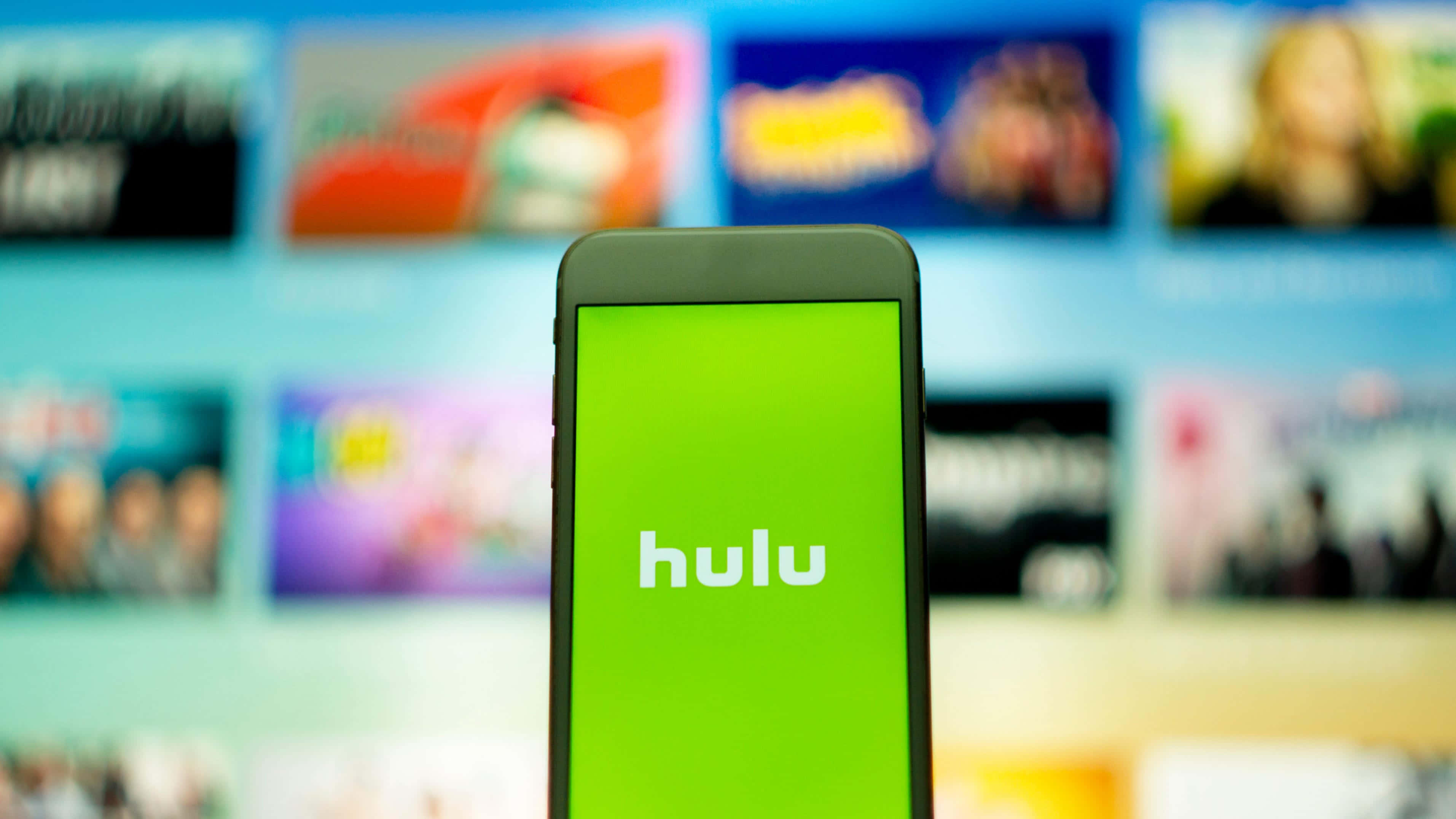 Hulu 桌布 壁紙 — 一部智慧型手機在亮綠色螢幕上以白色文字顯示 Hulu 標誌，背景是模糊的電視螢幕，顯示色彩繽紛的節目縮圖。