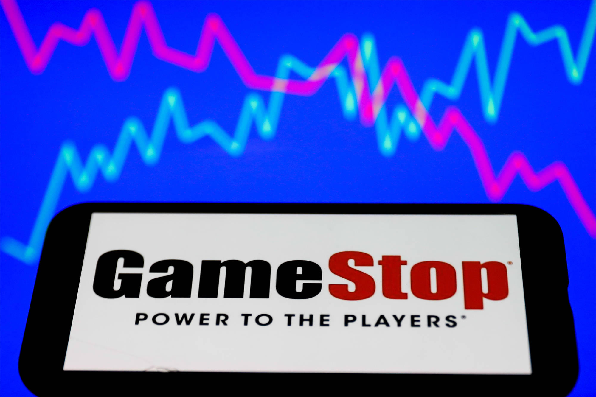 GameStop 桌布 壁紙 — 一部智慧型手機在充滿活力的藍色背景上顯示GameStop標誌，並帶有發光的粉紅色和青色股票市場圖表。