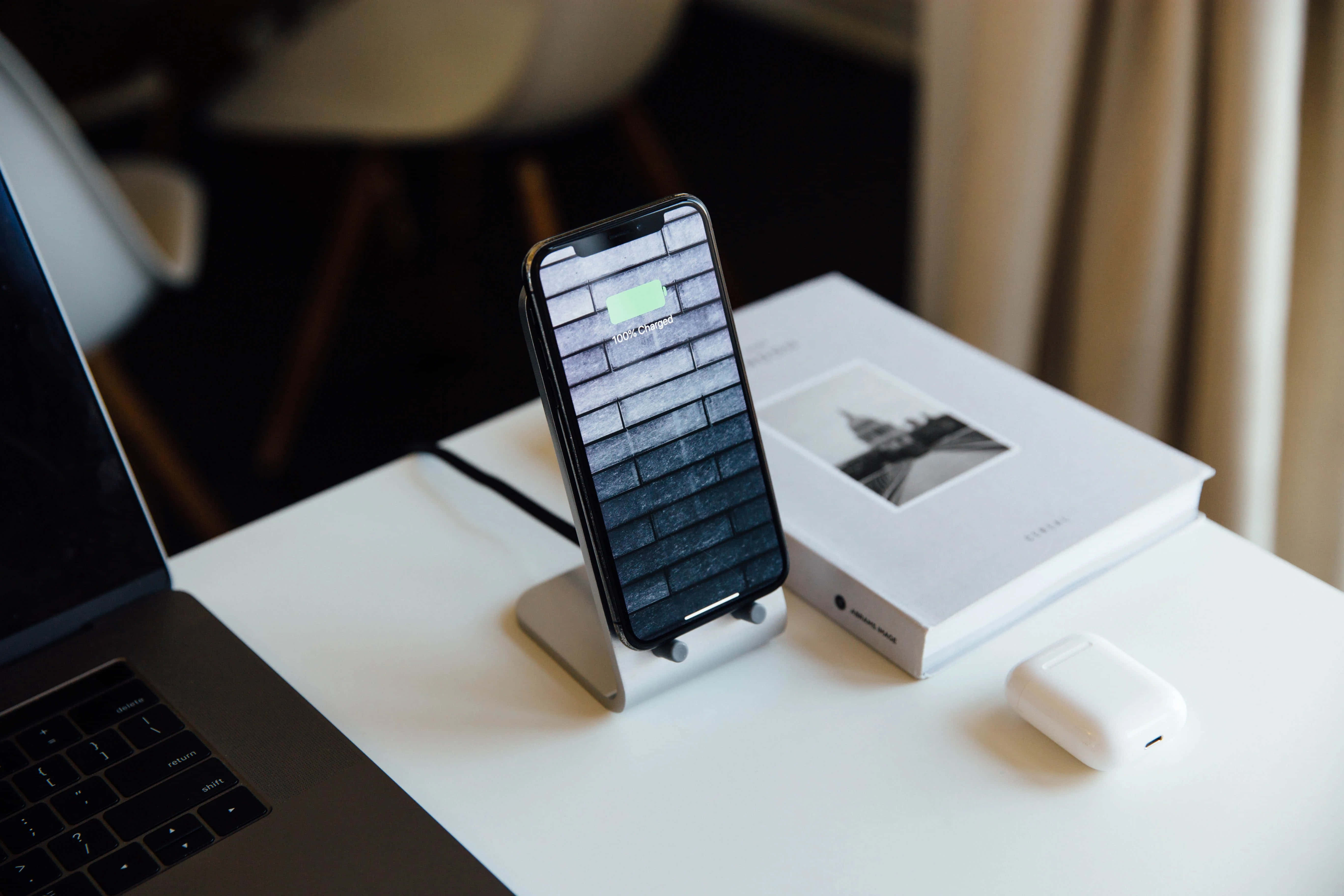 Wireless Charging 桌布 壁紙 — 一部智慧型手機在支架上充電，旁邊有一台筆記型電腦和AirPods，放在簡約的白色桌子上。