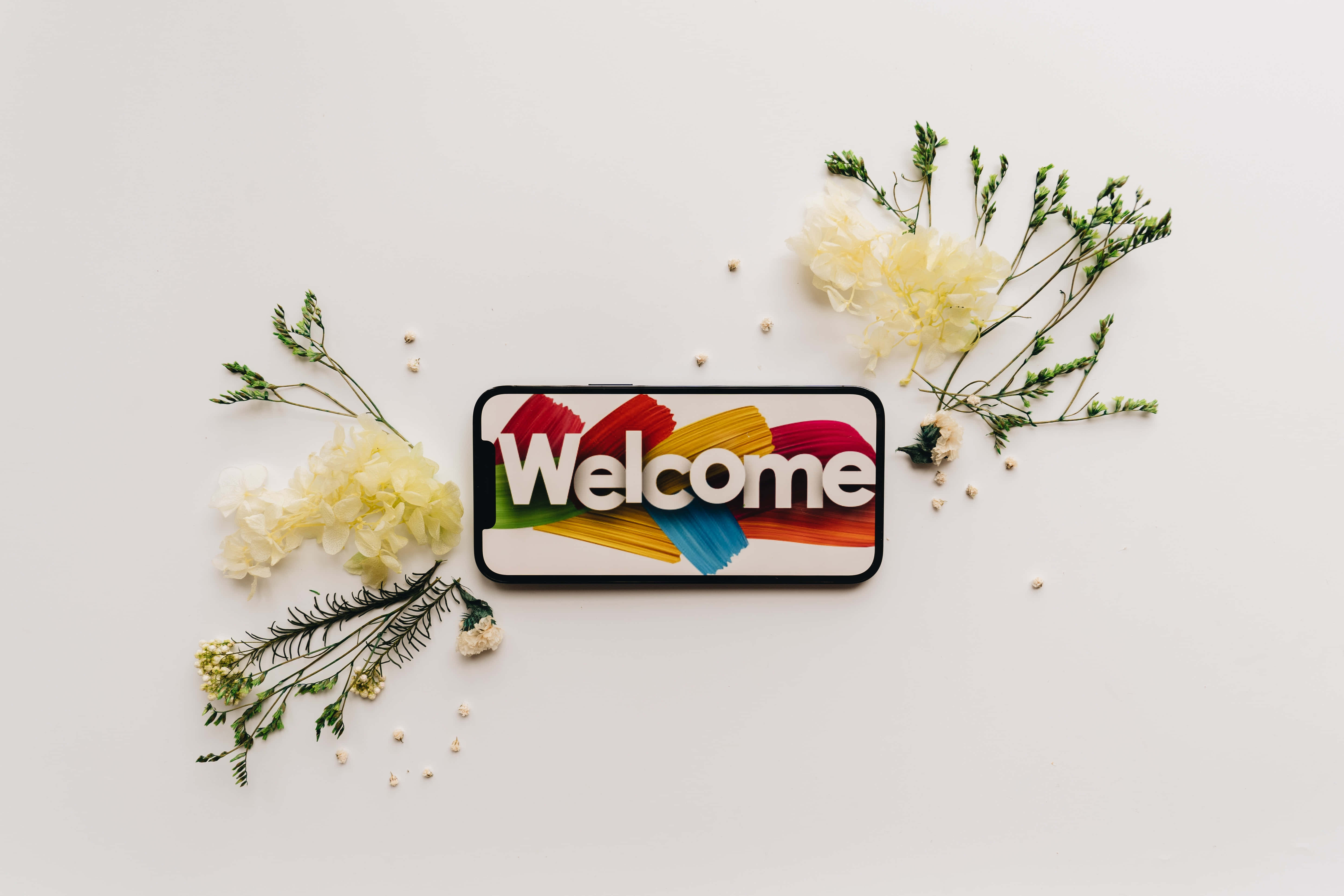 Welcome 桌布 壁紙 — 一部智慧型手機在白色表面上顯示色彩繽紛的「歡迎」圖案，周圍環繞著精緻的黃色花朵和綠色植物。
