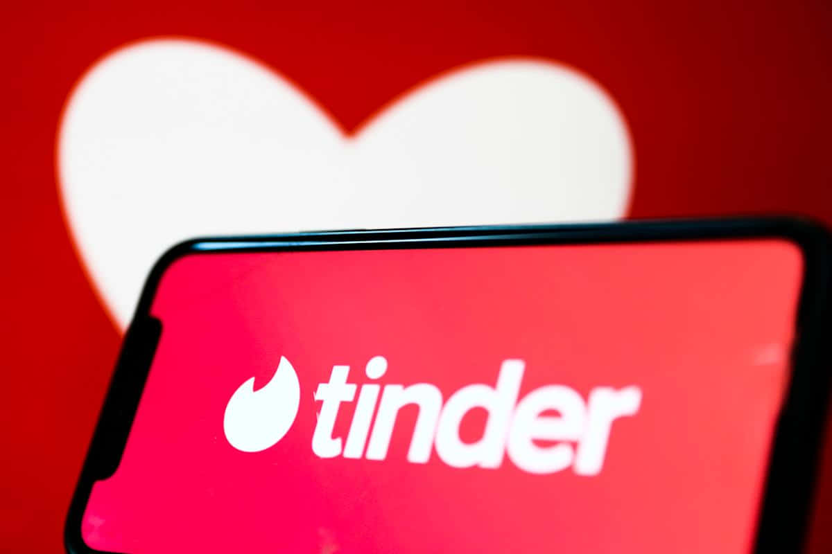 tinder 桌布 壁紙 — 一部智慧型手機在鮮紅色背景上顯示Tinder應用程式標誌，背後有一個柔軟的白色心形圖案。