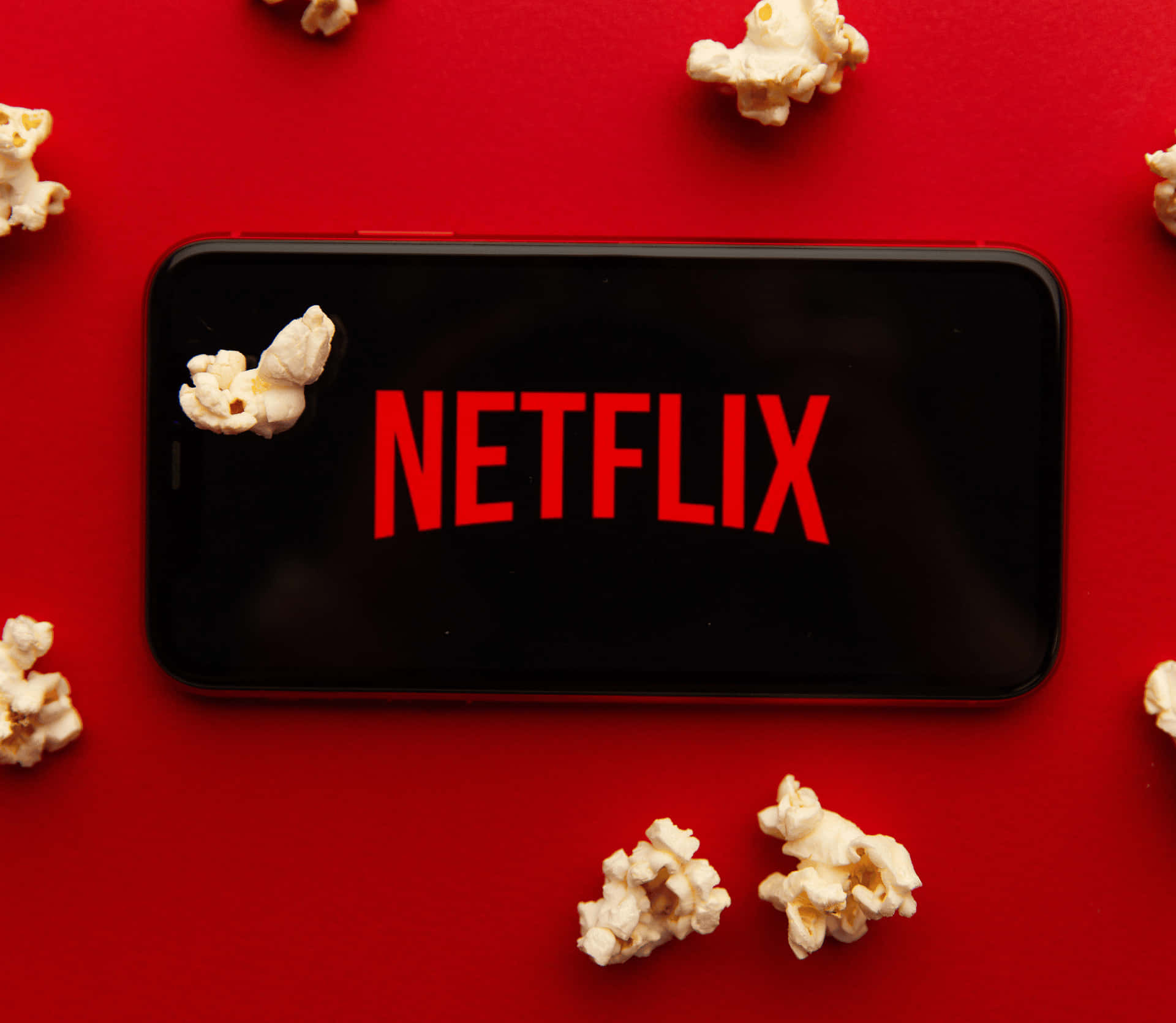 手機 桌布 壁紙 — 一部智慧型手機在鮮紅色表面上顯示Netflix標誌，周圍環繞著爆米花。