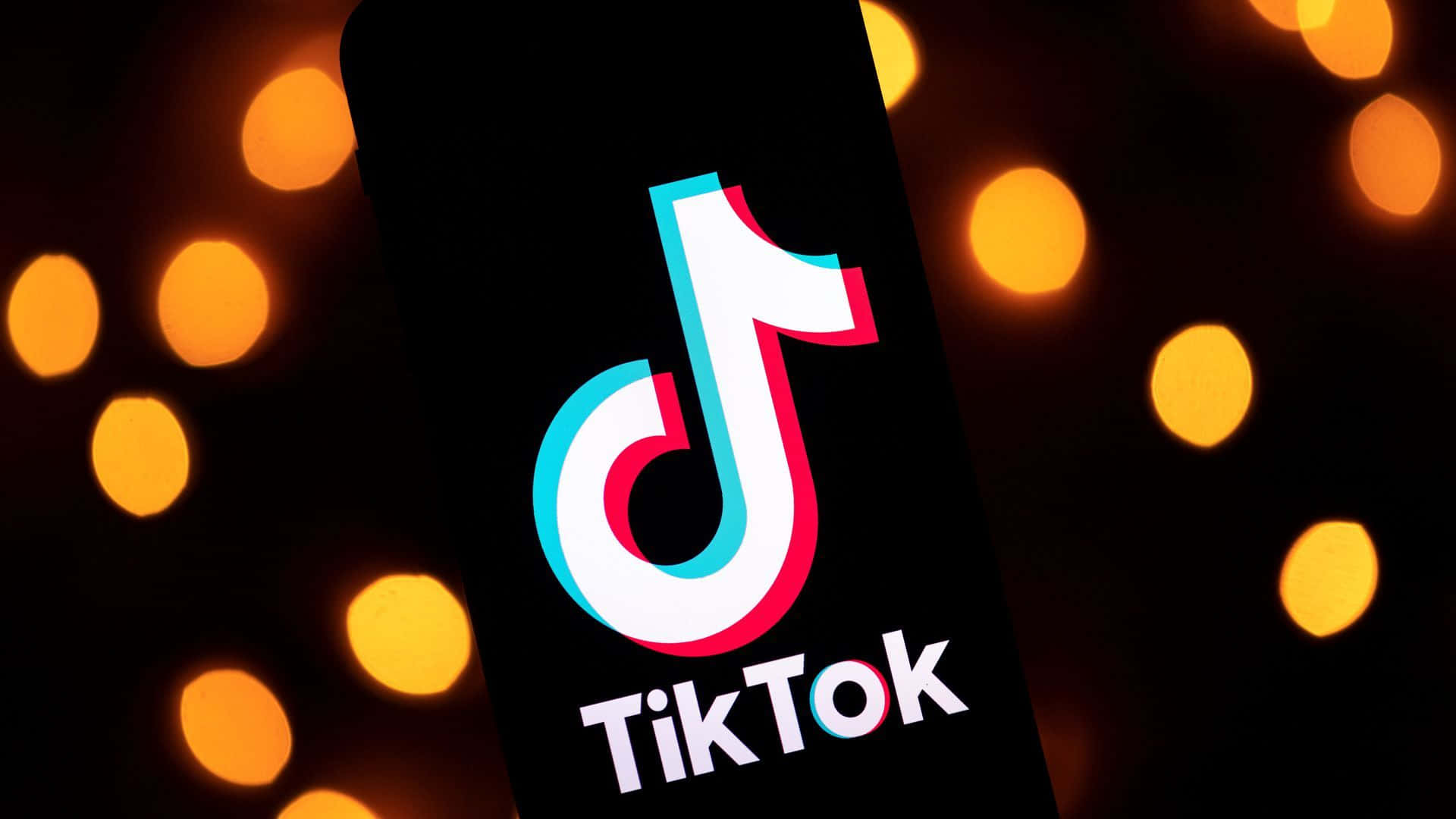 Bokeh 桌布 壁紙 — 一部智慧型手機在黑暗背景上顯示TikTok標誌，帶有溫暖的橙色散景燈光。