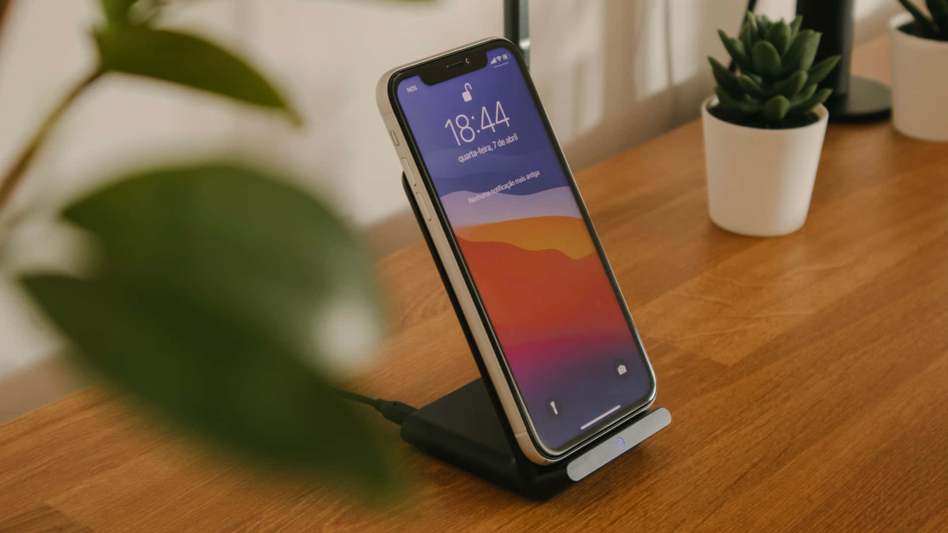 Wireless Charging 桌布 壁紙 — 一部智慧型手機放在無線充電座上，置於木桌旁，旁邊有盆栽植物，柔和自然光線。