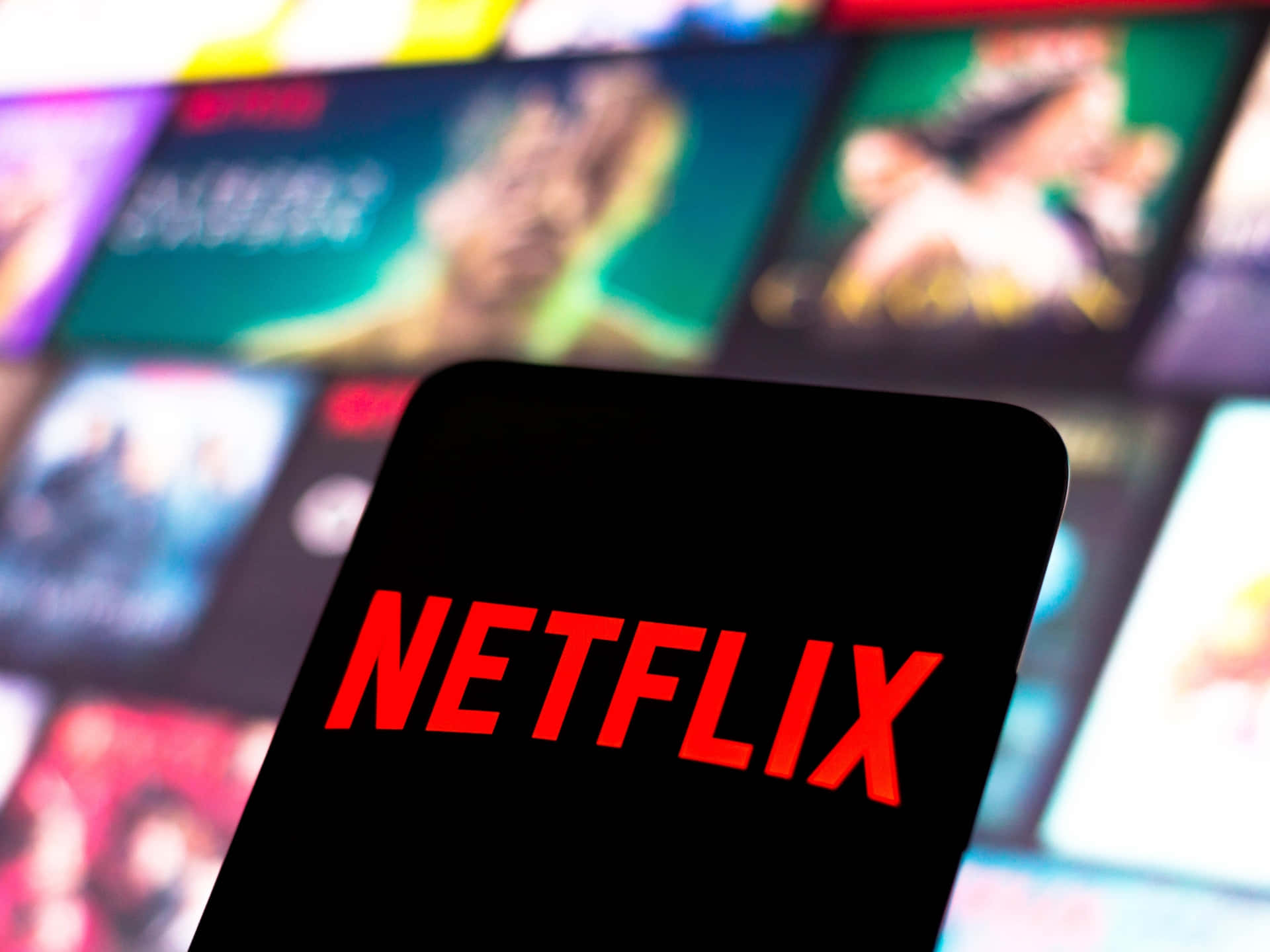 Netflix 桌布 壁紙 — 一部智慧型手機清晰顯示紅色Netflix標誌，背景是模糊的彩色串流縮圖。