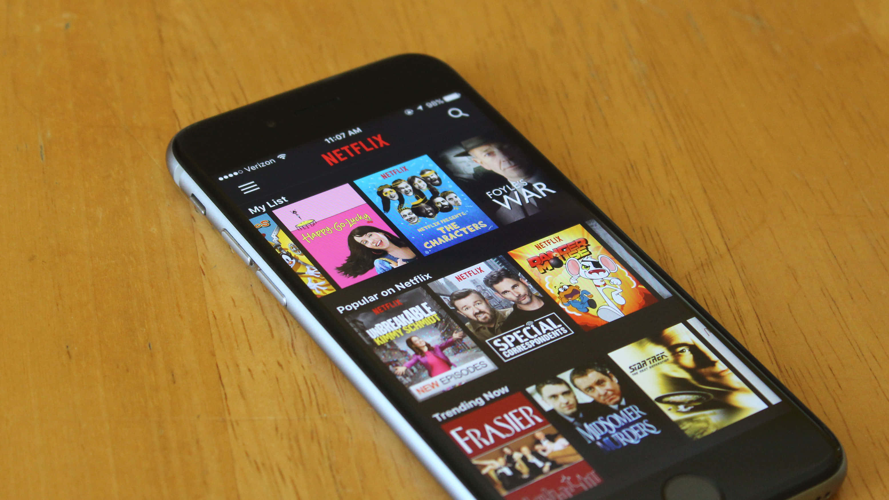 Netflix 桌布 壁紙 — 一部智慧型手機顯示Netflix應用程式，帶有色彩繽紛的節目縮圖，放置在溫暖的木質表面上。