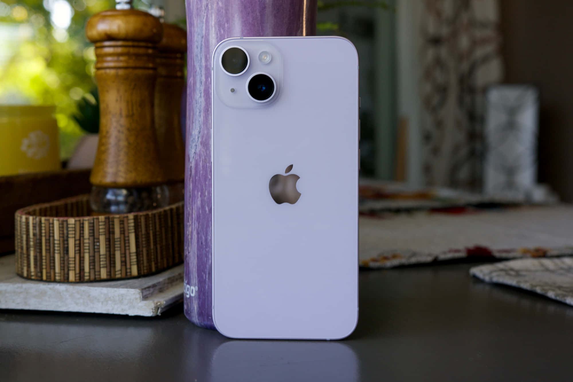 Apple Phone 桌布 壁紙 — 一部淺紫色iPhone 14直立放在一張深色桌子上，旁邊有一個木製胡椒磨和一個紫色花瓶，營造出舒適的廚房環境。