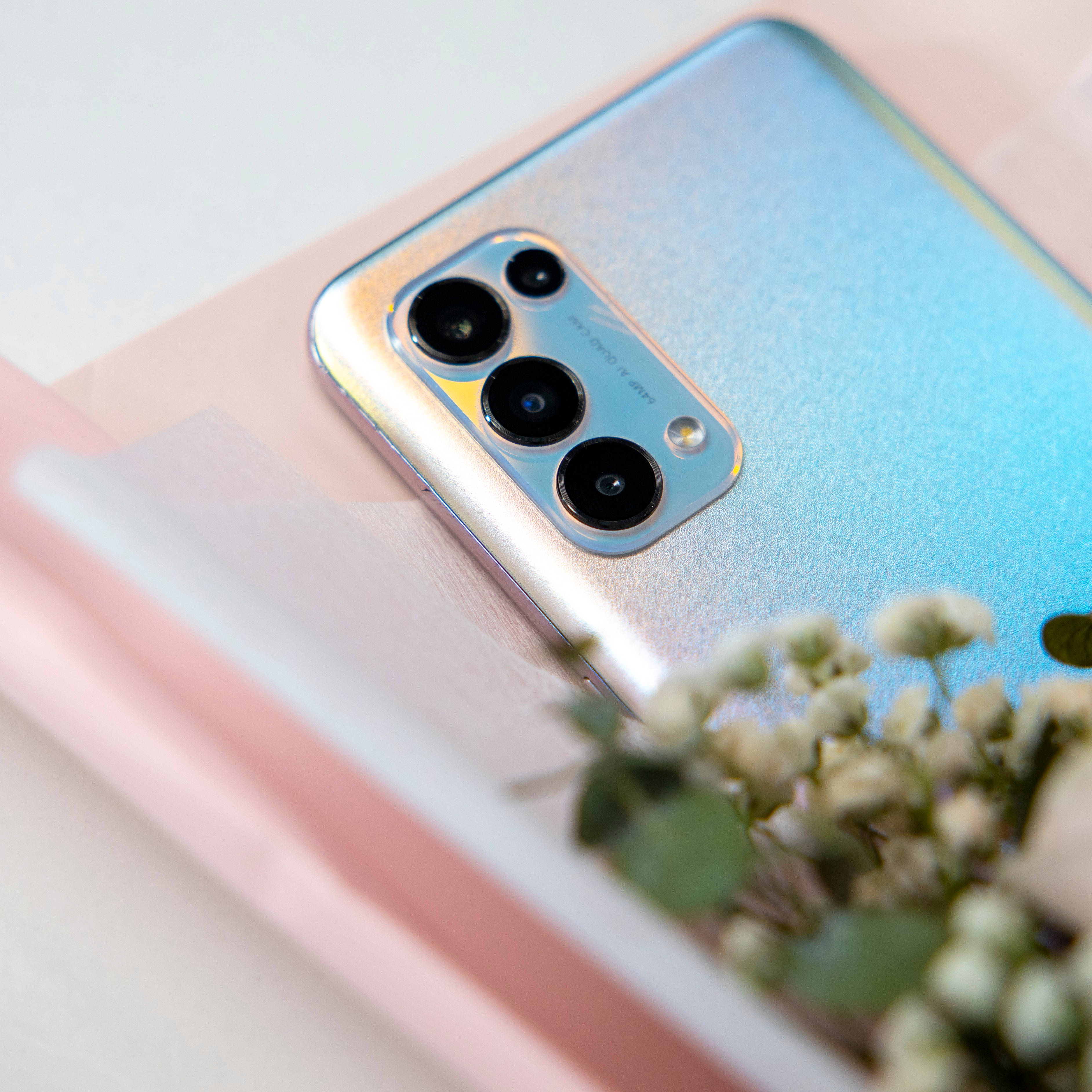 oppo 桌布 壁紙 — 一部漸層藍色智慧型手機的特寫，帶有四個鏡頭模組，放在一本粉紅色書本上，旁邊是精緻的白色花朵。