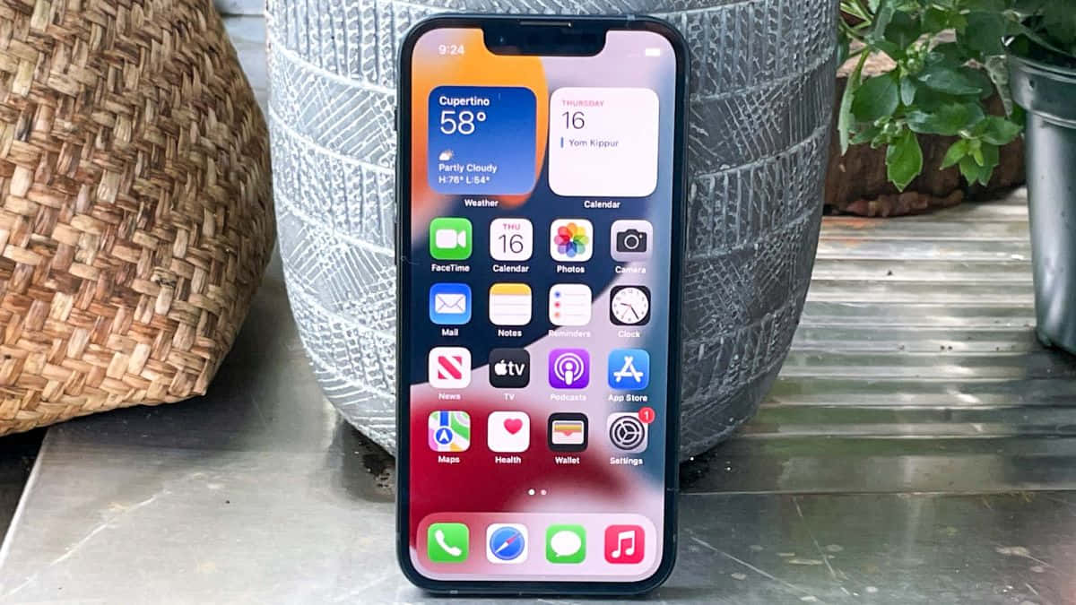 手機 桌布 壁紙 — 一部現代iPhone顯示色彩繽紛的主螢幕，帶有應用程式圖標和天氣資訊，放置在金屬表面上，旁邊有編織和陶瓷花盆。