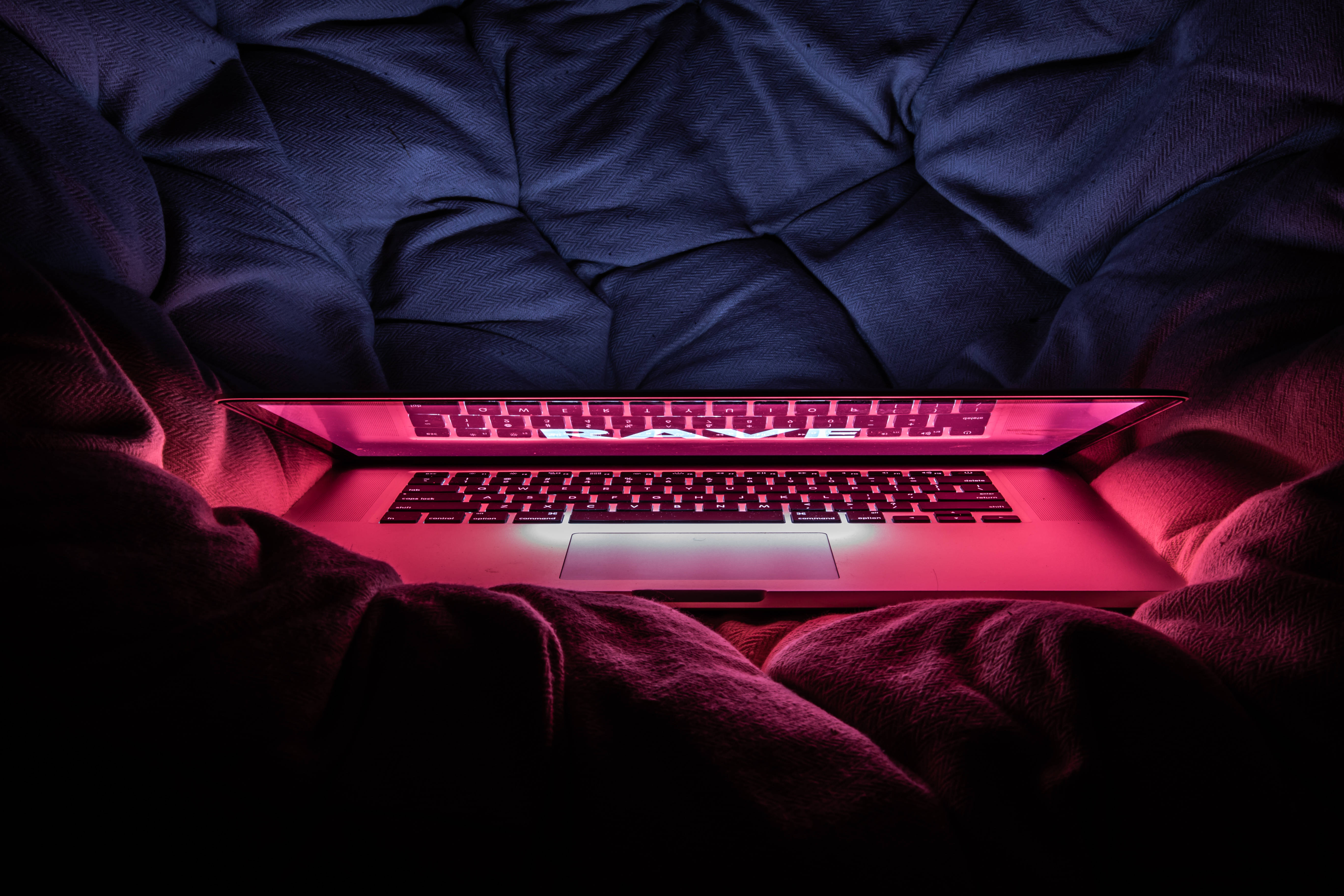 macbook pro 桌布 壁紙 — 一部發出粉紅色光的筆記型電腦放在昏暗房間中皺巴巴的深藍色床單上。